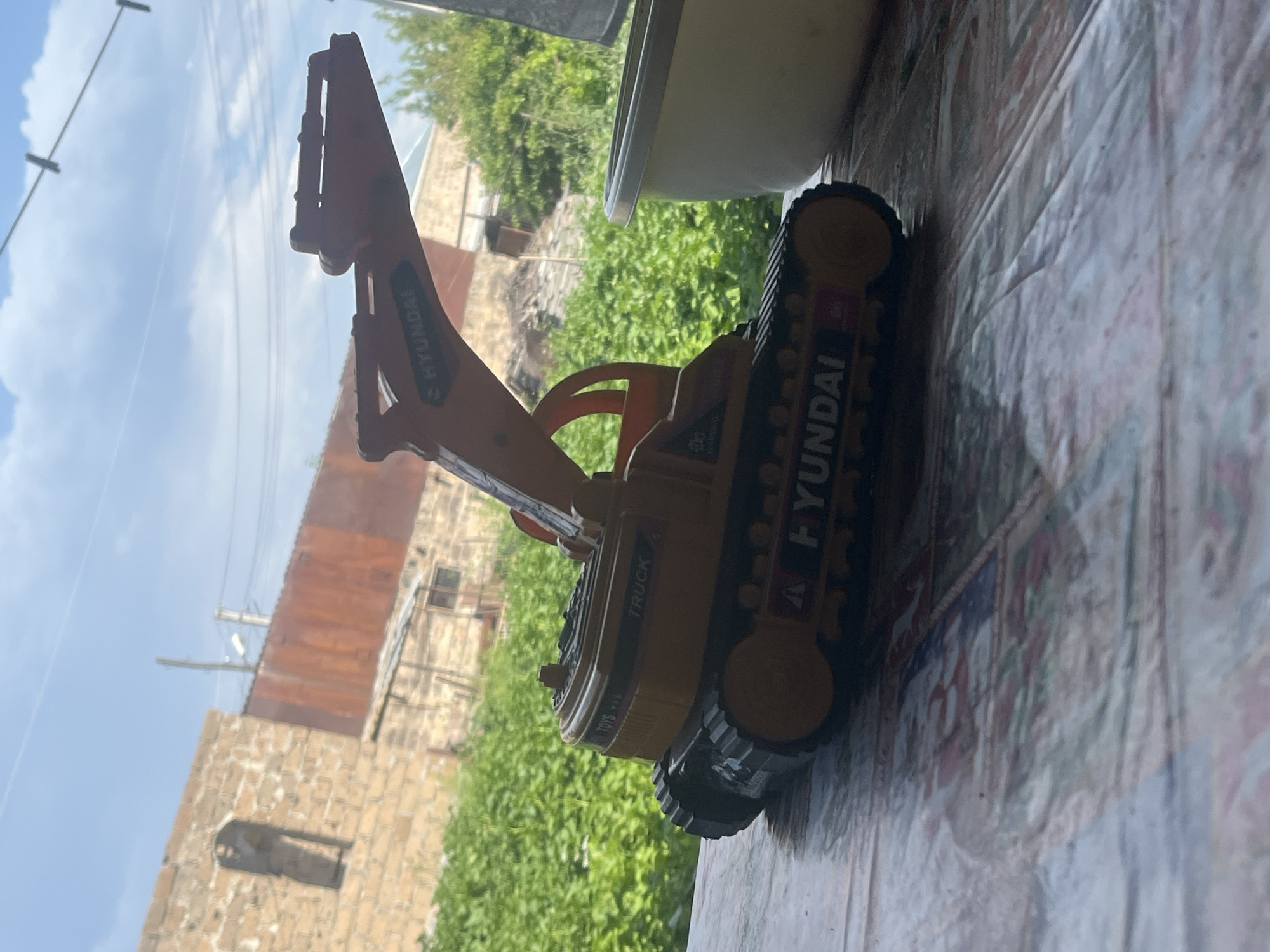 A toy excavator, yellow and orange, with "HYUNDAI" branding, sits on a patterned surface, possibly a tablecloth. The excavator is facing right, with its tracks and boom visible. In the background, out of focus, are green foliage, a brick building with an arched opening, and a rusted metal structure under a cloudy sky. The scene is viewed from a slightly elevated, close-up perspective. This image was taken in Jrarbi, Armenia.