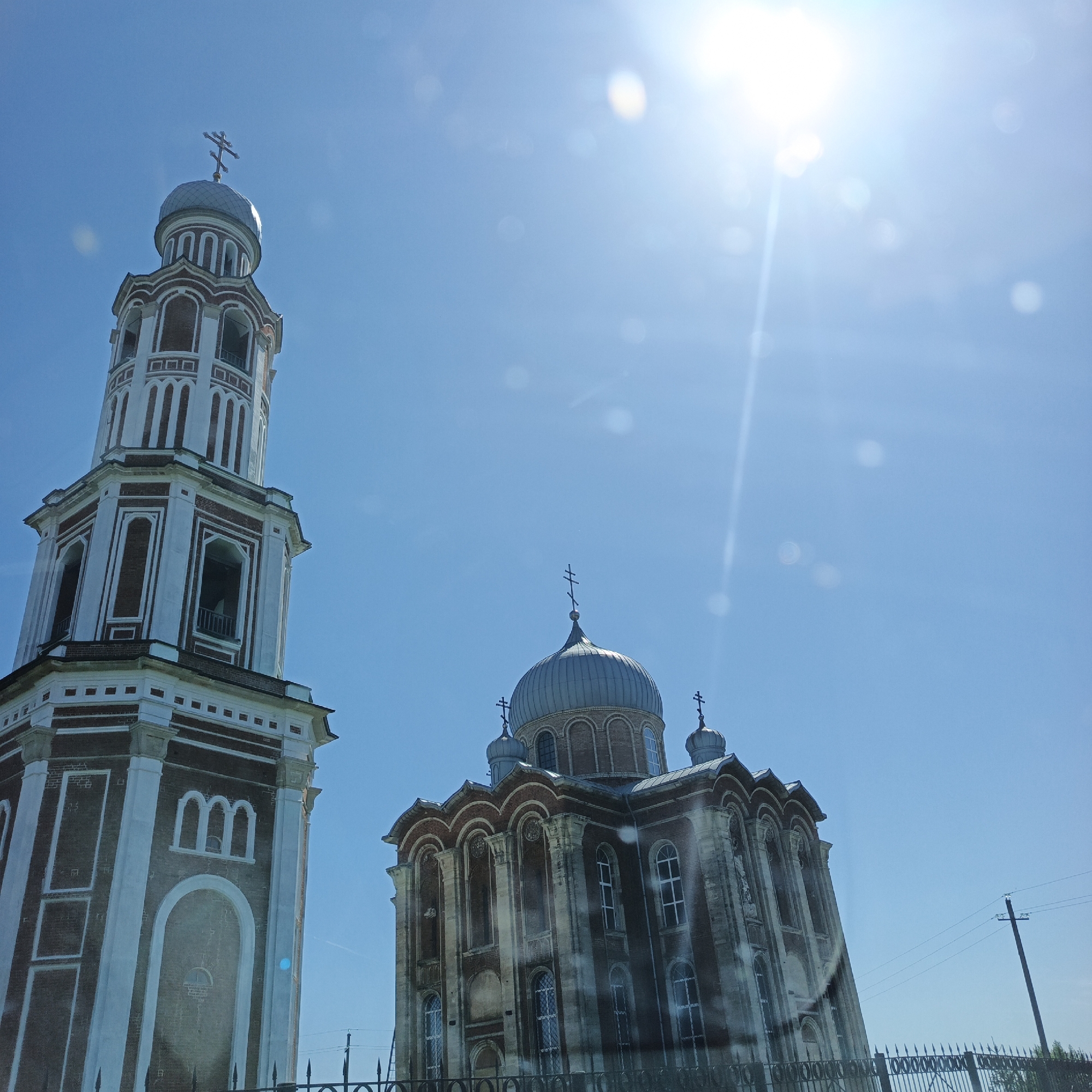 In Тугустемир, Russia, a bell tower and an Orthodox church stand against a bright blue sky. The bell tower, on the left, is tall and square, with red brick and white trim. It features arched openings with bells visible within. A silver dome with a cross crowns the tower. To the right, the church has a large central dome, also silver and topped with a cross, and smaller domes on either side. The church's facade is a mix of red brick and weathered stone, with arched windows. The sun is prominently visible in the upper right corner, casting a bright glare and lens flares across the sky, obscuring some details of the buildings. A utility pole with wires is visible in the background on the right, and a black metal fence runs across the bottom of the image. The scene appears to be an exterior view on a sunny day.