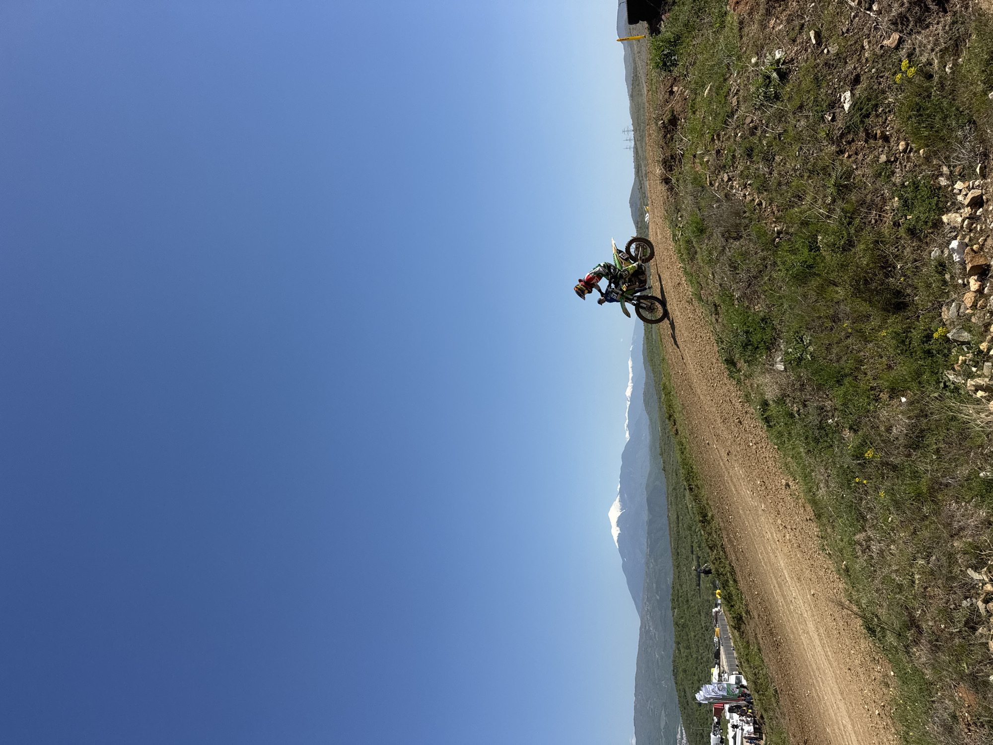 A dirt biker is on a dirt track in Selishte, Bulgaria. The biker is wearing a green and red suit with a helmet. The biker is riding on a green dirt bike. The track is on the side of a hill with mountains in the background. The sky is blue. There are people and tents at the bottom of the hill.