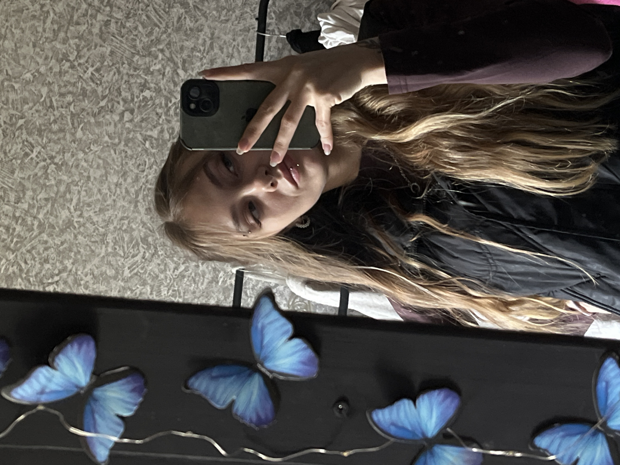 A young woman is taking a selfie in a mirror. The mirror is decorated with blue butterfly lights strung along the bottom. The woman has blonde hair and piercings on her face. She is holding an iPhone. The environment is an indoor space. No location is confirmed, or Фоки, Russia.