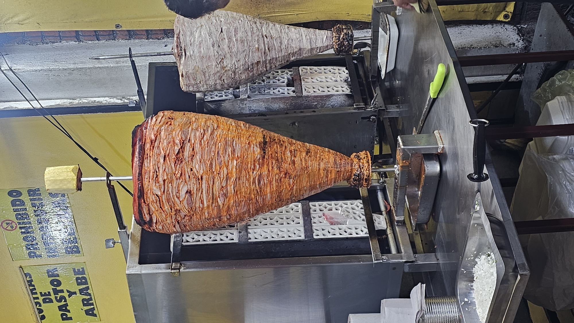 The image shows two vertical rotating spits of meat, likely for tacos al pastor or shawarma, in a food preparation area. The spit in the foreground is cooked and reddish-orange in color, while the one in the background is lighter, raw meat. The spits are mounted on a metal stand with white, decorative brick-like heat deflectors. A yellow sign with Spanish text, including "PROHIBIDO" (prohibited) and "TAQUINAS DE PASTOR Y ARABE" (Tacos al Pastor and Arabe), is visible to the left of the foreground spit. A knife with a green handle and a metal blade is visible on the right. A pile of what appears to be chopped onions is near the edge of the counter. Based on the sign, the location is likely a street food stand in San Bernardino Tlaxcalancingo, Mexico.