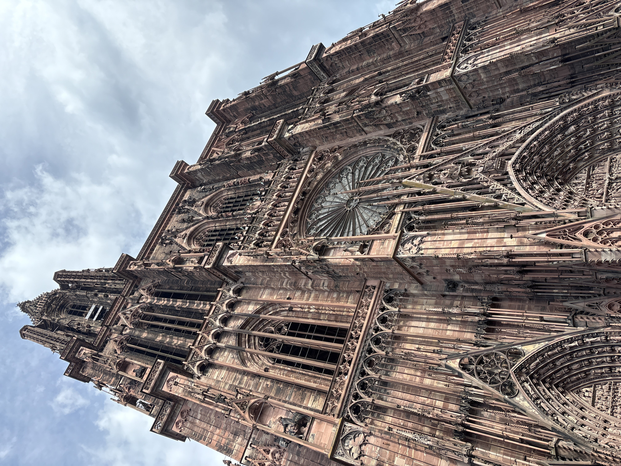 The image shows a worm's-eye view of Strasbourg Cathedral, a Gothic masterpiece located in Strasbourg, France. The cathedral is made of pink sandstone, and its intricate façade features elaborate carvings, sculptures, and rose windows. The architecture exhibits ornate stonework, including arches, spires, and statues. The sky is cloudy and overcast. There are no people visible in the shot.