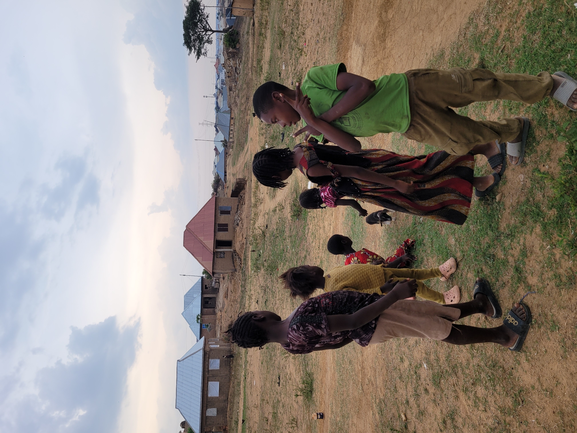 The image depicts five children in a grassy area in Barakin Akawo, Nigeria. The children are standing and sitting. One boy is wearing a green shirt and khaki pants, and has his hands up near his face. The children are interacting with each other. In the background are several single-story buildings with roofs of varying colors. The sky is partly cloudy. A tree is visible in the far background. The ground is a mix of bare earth and grass.