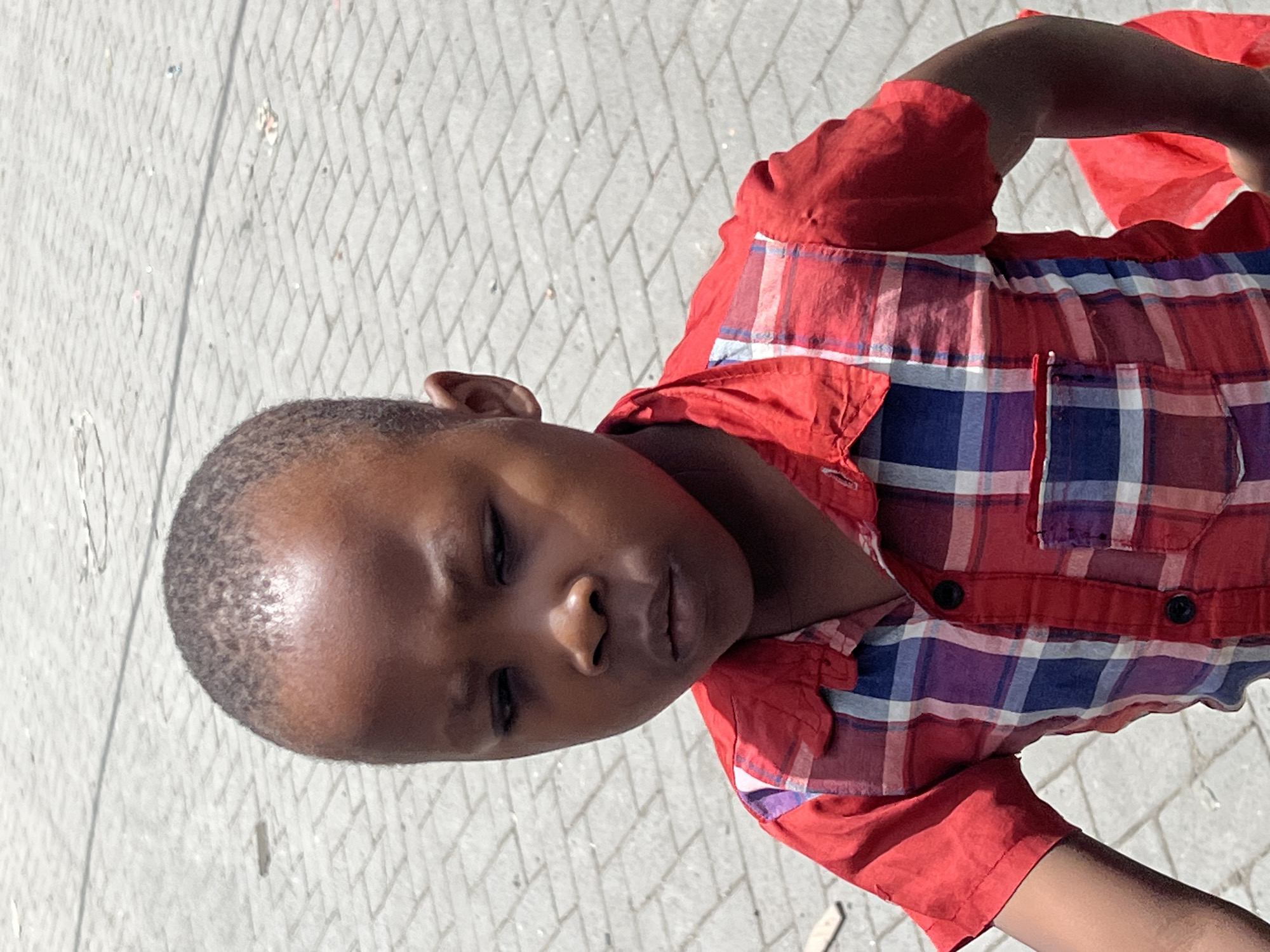 A close-up shows a young Black boy wearing a red plaid shirt with blue and white accents. The shirt has a pocket on the front and is buttoned. He appears to be outside, standing on a paved surface of interconnected geometric shapes, likely in a public area. The light is bright, suggesting it is daytime. The location is not evident from this single image. Without additional contextual information, it is impossible to determine if this image was taken in Nairobi, Kenya, or any other location.
