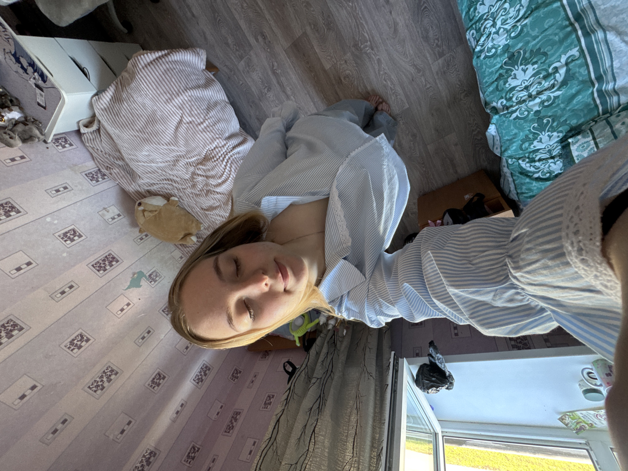 A top-down shot shows a blonde woman lying in a room. She wears a blue and white striped shirt. The floor is wood-look laminate in some areas and patterned tile in others. Near her head are curtains. A window with a white frame is visible with greenery outside. Other objects include a white cabinet, a striped blanket, a stuffed animal, and a patterned bedspread. The location is likely an interior space in Perm, Russia, based on environmental cues that are typical to the country.