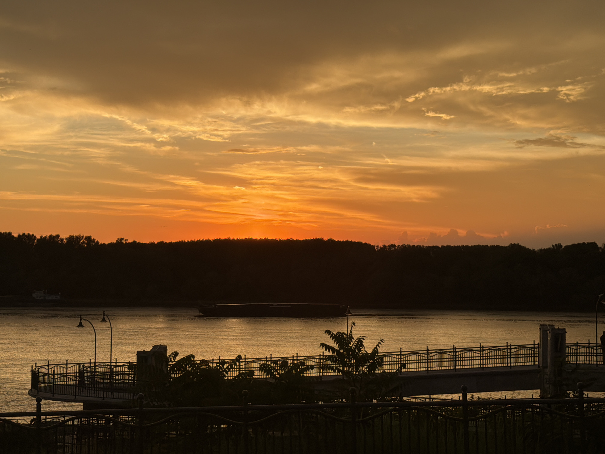 The image depicts a sunset over the Danube River in Ruse, Bulgaria. The sky is filled with vibrant orange and yellow hues, with scattered clouds. A dark silhouette of trees lines the riverbank. In the foreground, a promenade with a decorative black metal railing is visible, along with streetlights. A barge is seen sailing on the river. The overall scene is tranquil and picturesque.