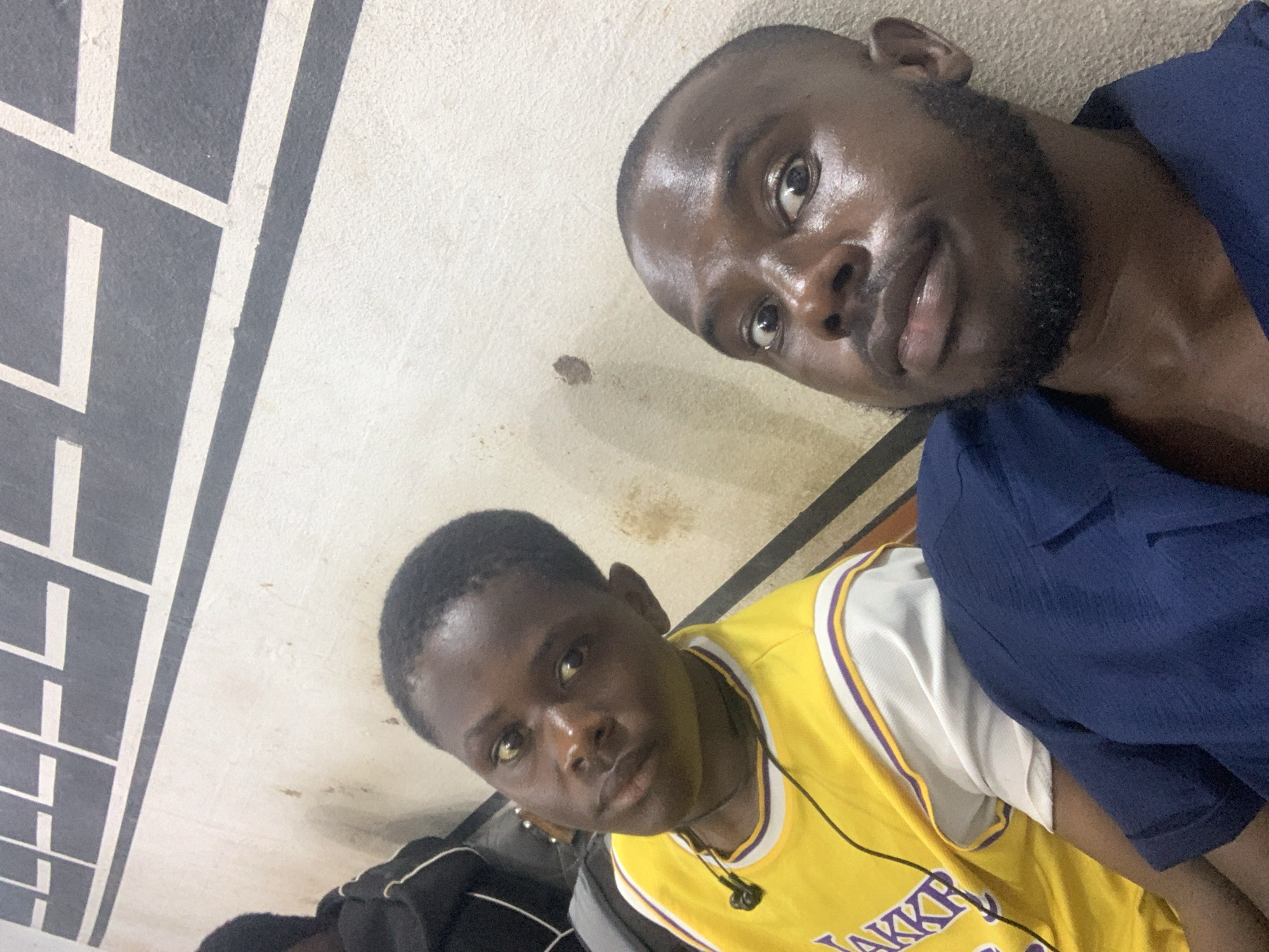 Two men pose for a selfie in an indoor location, possibly in Jos, Nigeria, judging by the men's appearances. The man on the left wears a yellow Lakers jersey and black earbuds. The man on the right has a shaved head, a beard, and is wearing a blue shirt. The background includes a patterned wall with horizontal lines. There are spots on the wall. The floor appears to be a basketball court.