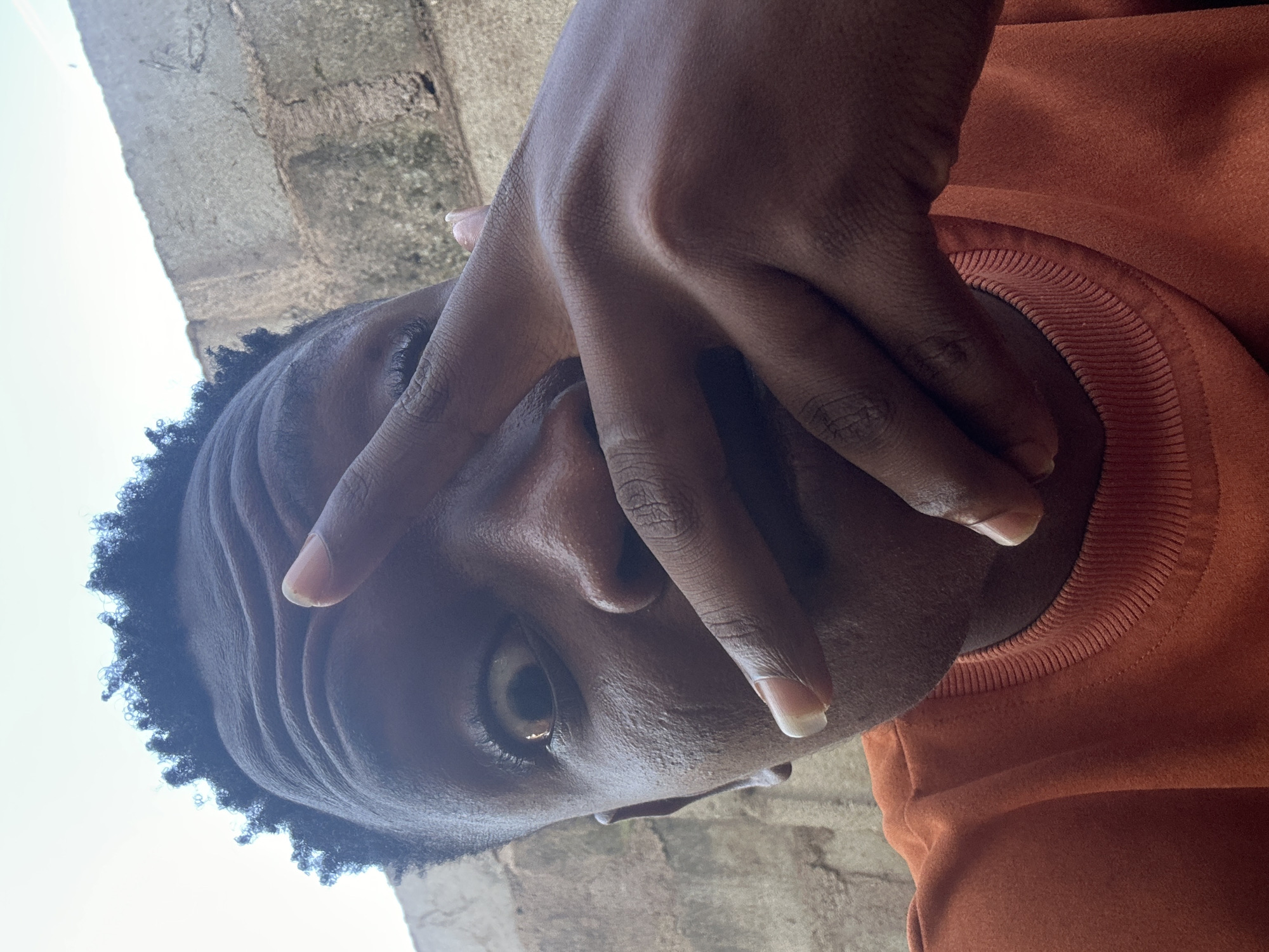 This is a close-up, low-angle shot of a young Black person, likely male, in what appears to be an outdoor setting in Ikorodu, Nigeria. The subject's head is tilted upwards, and their right hand is held up, covering part of their face, with fingers splayed across their forehead and eye. Only one eye is clearly visible, looking directly at the camera with an intense gaze. The subject has dark skin, short, dark, curly hair, and is wearing an orange sweatshirt with a ribbed neckline.
The background consists of a textured concrete wall on the left and a bright, overcast sky on the right. The lighting suggests it is daytime, possibly late morning or early afternoon, given the brightness of the sky. The overall impression is one of a candid, perhaps playful or curious moment captured spontaneously. The subject's expression is difficult to fully ascertain due to the hand partially obscuring their face, but the visible eye conveys a direct and engaging look. There is no visible text in the image.