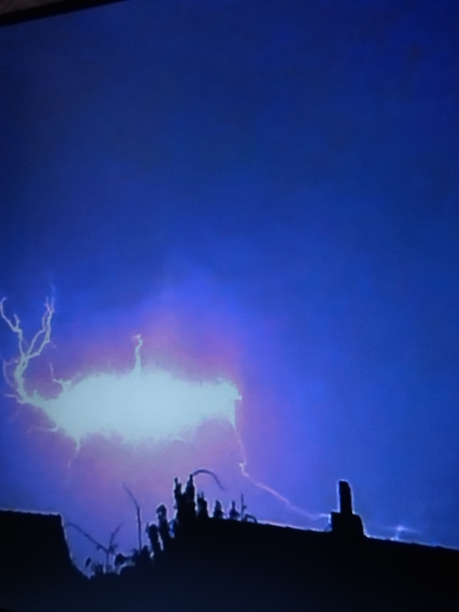 A silhouette of a town is visible against a stormy, purple-hued sky. Lightning flashes illuminate the clouds, with a bright, wide strike dominating the center left of the frame. The dark outlines of buildings and trees are discernible against the dramatic sky. The scene appears to be a nighttime thunderstorm over an urban or semi-urban area.