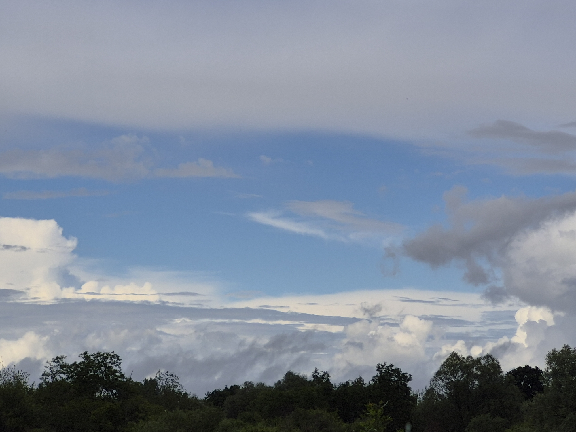 The image shows a landscape with trees in the foreground and a cloudy sky above. The trees are dark green and appear to be dense forest. The sky is a mix of blue and grey clouds. Some of the clouds are white and fluffy, while others are dark and menacing. The clouds are arranged in layers, with a band of blue sky visible in the middle. The overall impression is one of a natural, unpopulated landscape. There are no people or man-made objects visible in the image. The location is in Sooblase, Estonia.