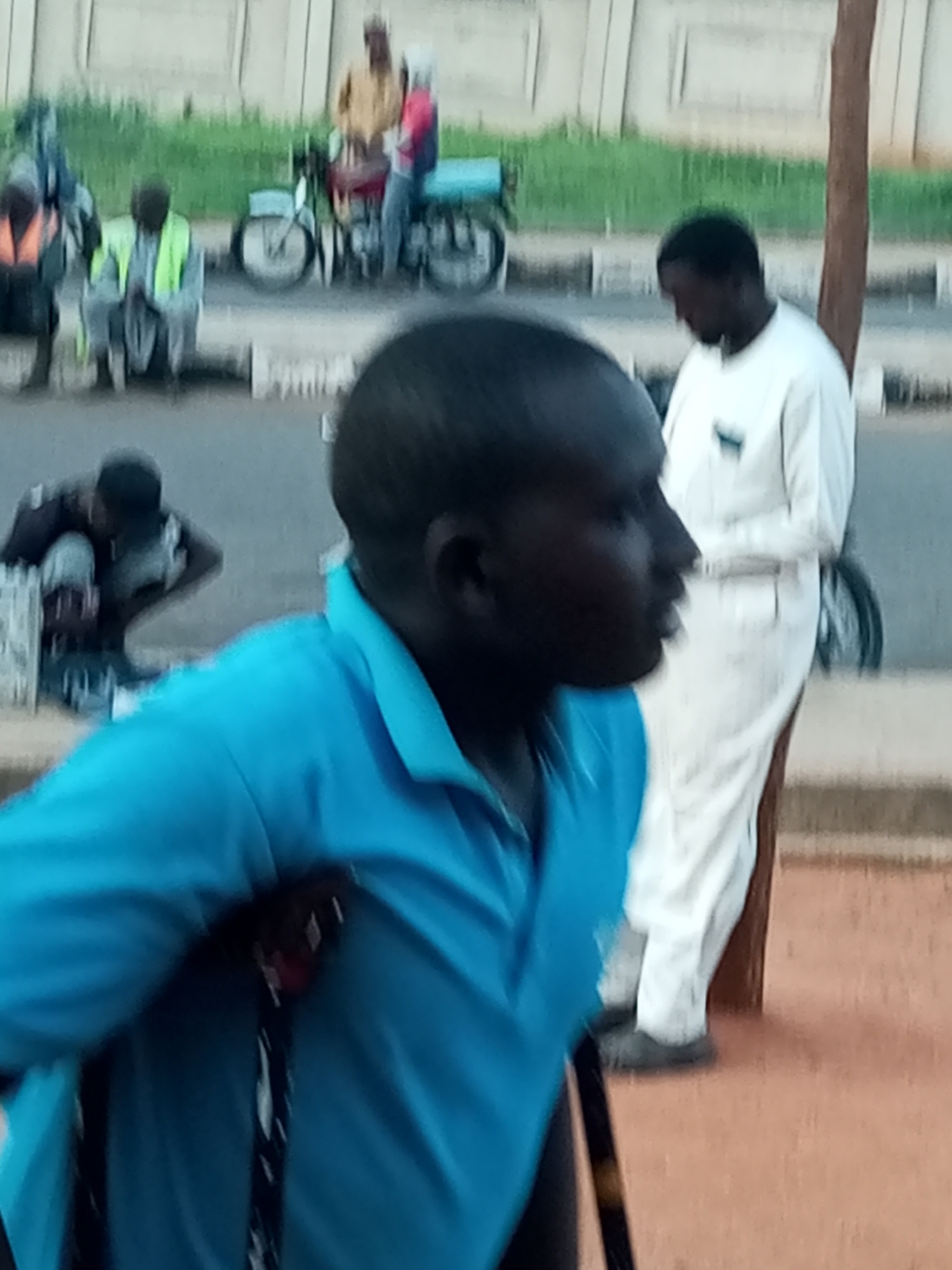 A medium shot captures a group of people near a roadside in Gombe, Nigeria. In the foreground, a person wearing a blue collared shirt, possibly a polo shirt, is visible from the chest up, facing to the right and slightly downward. Their dark skin and short hair are discernible. They are holding a cane or walking stick, partially obscuring the left side of their chest.
In the mid-ground, another individual stands, wearing a long white robe and pants. They are facing away from the camera and looking downwards. To the left and further back, several other individuals are positioned. One person is seated, wearing a bright green vest over a grey shirt and pants. Another individual is standing behind them, wearing a dark shirt with an orange vest over it.
Further in the background, a motorcycle with a sidecar is visible. Two individuals are seated on or near the motorcycle. One is wearing a tan or yellow shirt and the other a red and blue garment. A thin tree trunk is positioned vertically on the right side of the frame. The environment appears to be a roadside with a light-colored wall or building in the distance and a grassy strip between the road and the wall. The ground in the foreground is a reddish-brown color, resembling dirt or paving. The lighting suggests it might be late afternoon or early evening, with soft shadows.