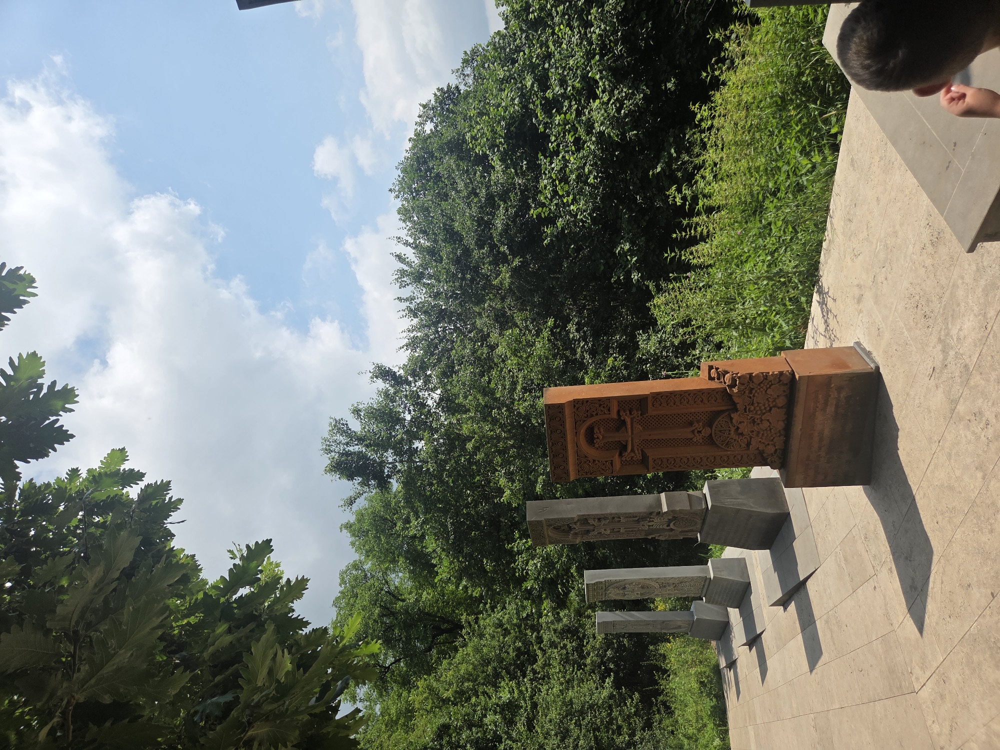 The image shows a row of khachkars (stone crosses) in Teghut, Armenia. The khachkars are carved with intricate designs and stand on a paved courtyard. In the background, lush green trees form a dense forest, with a bright blue sky and white clouds overhead. Sunlight casts long shadows on the courtyard, indicating it is a sunny day. A person's head and shoulder are visible in the lower right corner of the image, suggesting they are observing the khachkars.