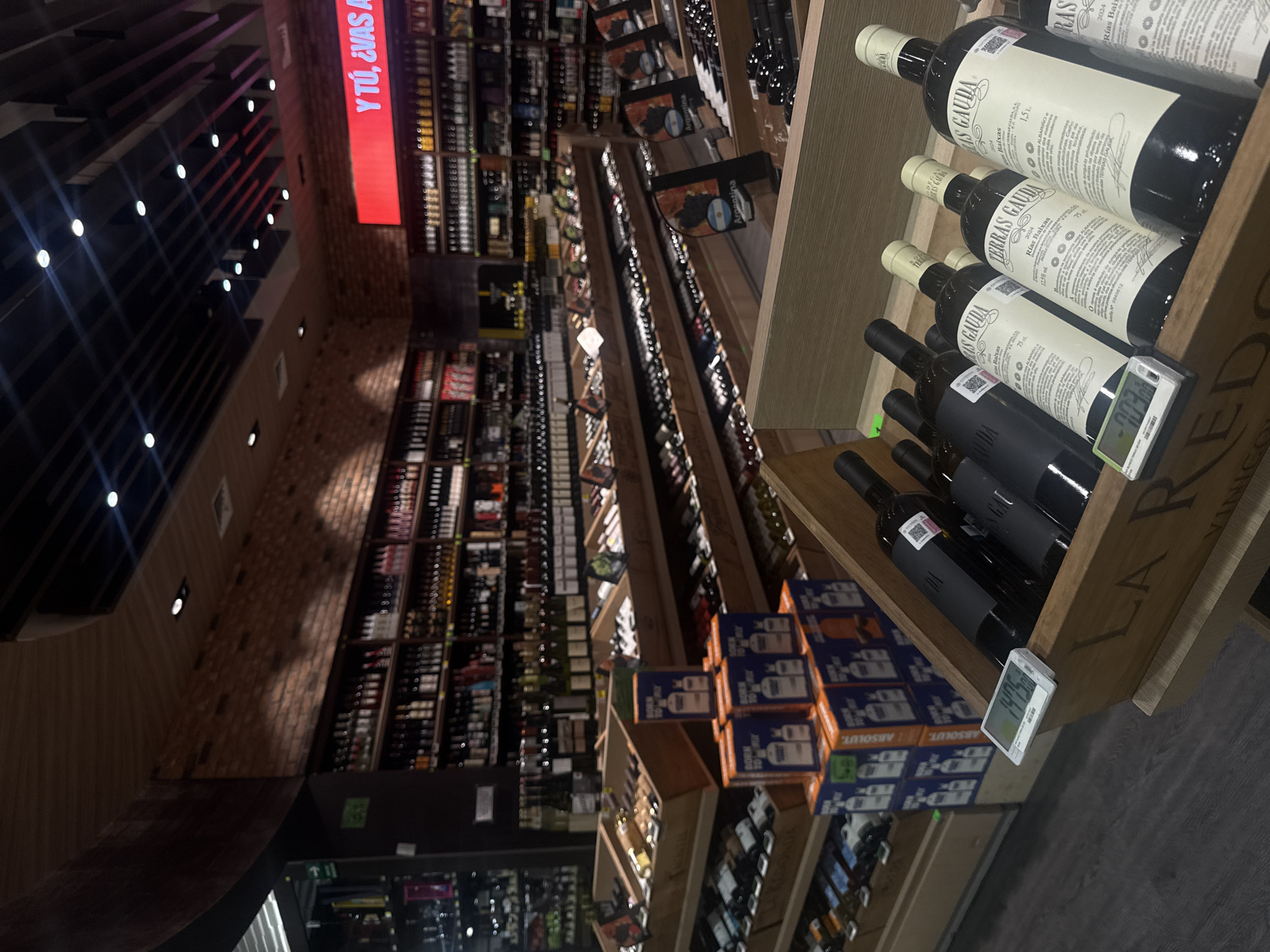 A high-angle, indoor shot showcases a well-stocked wine and spirits aisle, situated in an unknown location within an unknown city. The focus is on a wooden shelf unit containing bottles of "Terras Gauda Rias Baixas" wine, alongside boxes of "Absolut" vodka. Price tags are visible on some bottles, indicating potential costs. A large red sign with white text reading "Y TÚ, ¿VAS A" is visible on the left side. The general ambiance of the space implies a liquor store or a high-end market setting.