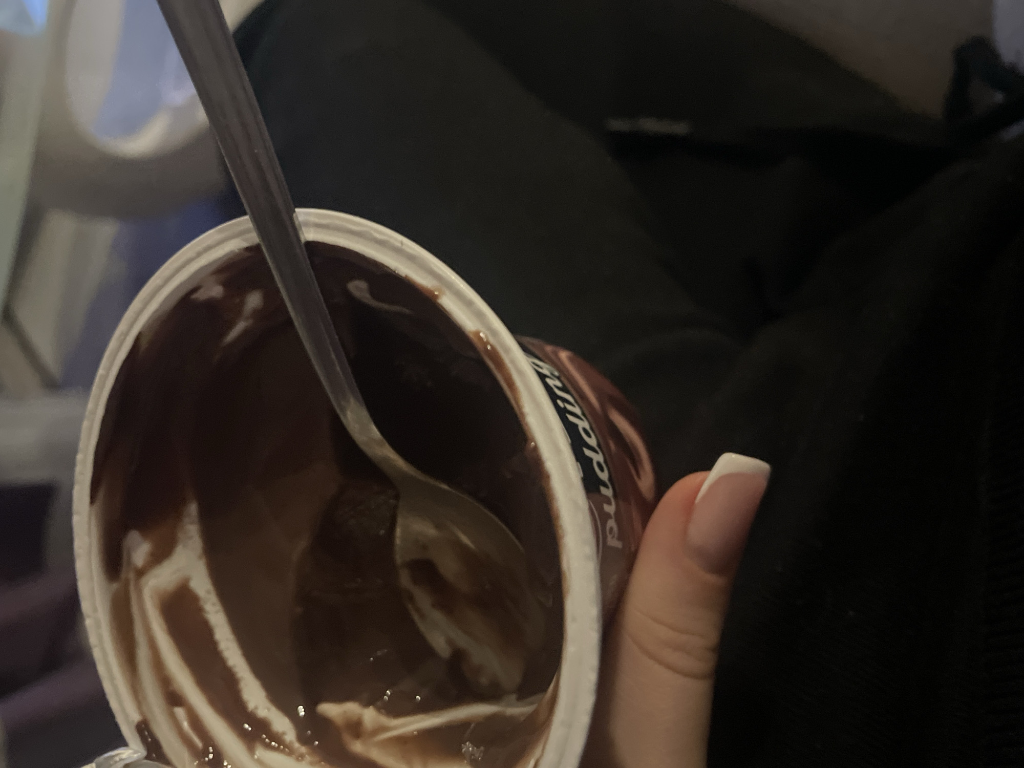 A close-up shot shows a partially consumed cup of chocolate pudding, held by a hand with a French manicure. A spoon is inside the cup, used to scoop out the pudding. The background is blurry, appearing to be a dark-colored surface, possibly clothing. The environment suggests an indoor setting. There is no data to place the location as Sofia, Bulgaria.