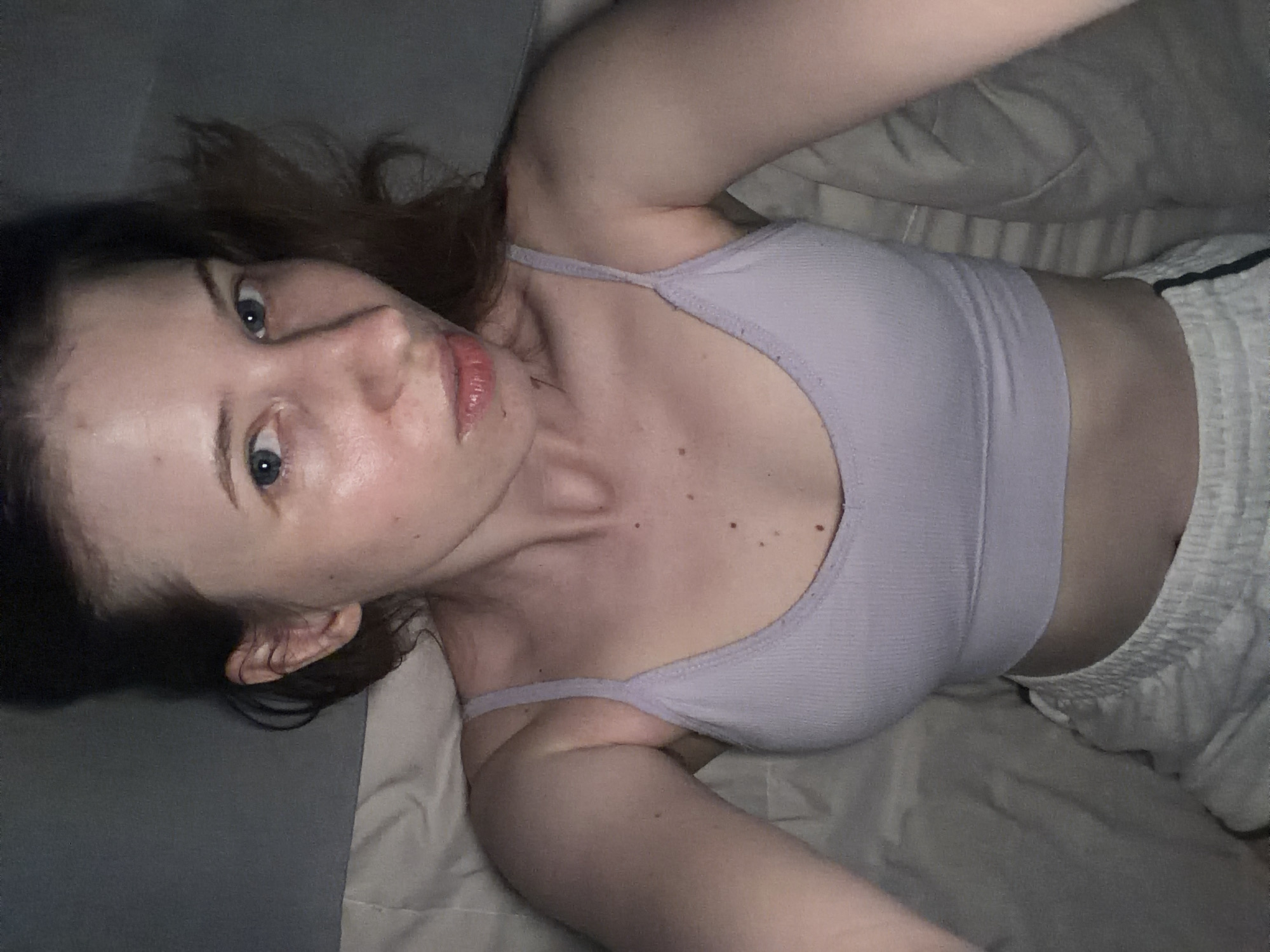 A person is lying on a bed in an upside-down orientation. They are wearing a light purple tank top and white shorts with black stripes. Their arms are outstretched above their head, and their head is tilted back, with their face turned towards the camera. The person has blue eyes and pink lips. The background consists of a bed with light-colored sheets. The lighting is dim, creating shadows on the person's face and body.