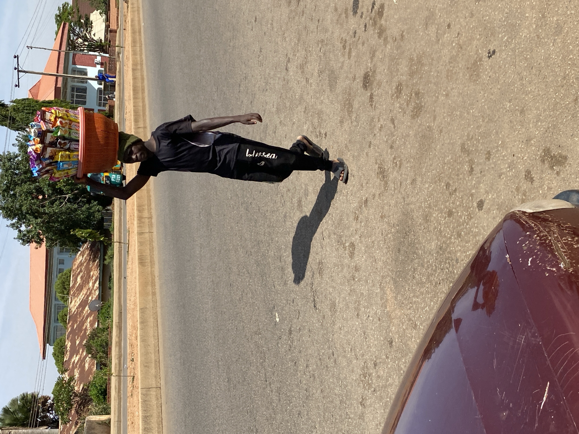 A man in Bukuru, Nigeria is carrying a large, orange basket filled with various goods on his head. He is walking on a paved road, and the edge of a maroon vehicle is visible in the foreground. The man is wearing a dark-colored shirt and pants with the word "blessed" on the pants leg and sandals. The background includes buildings, trees, and a light blue sky.