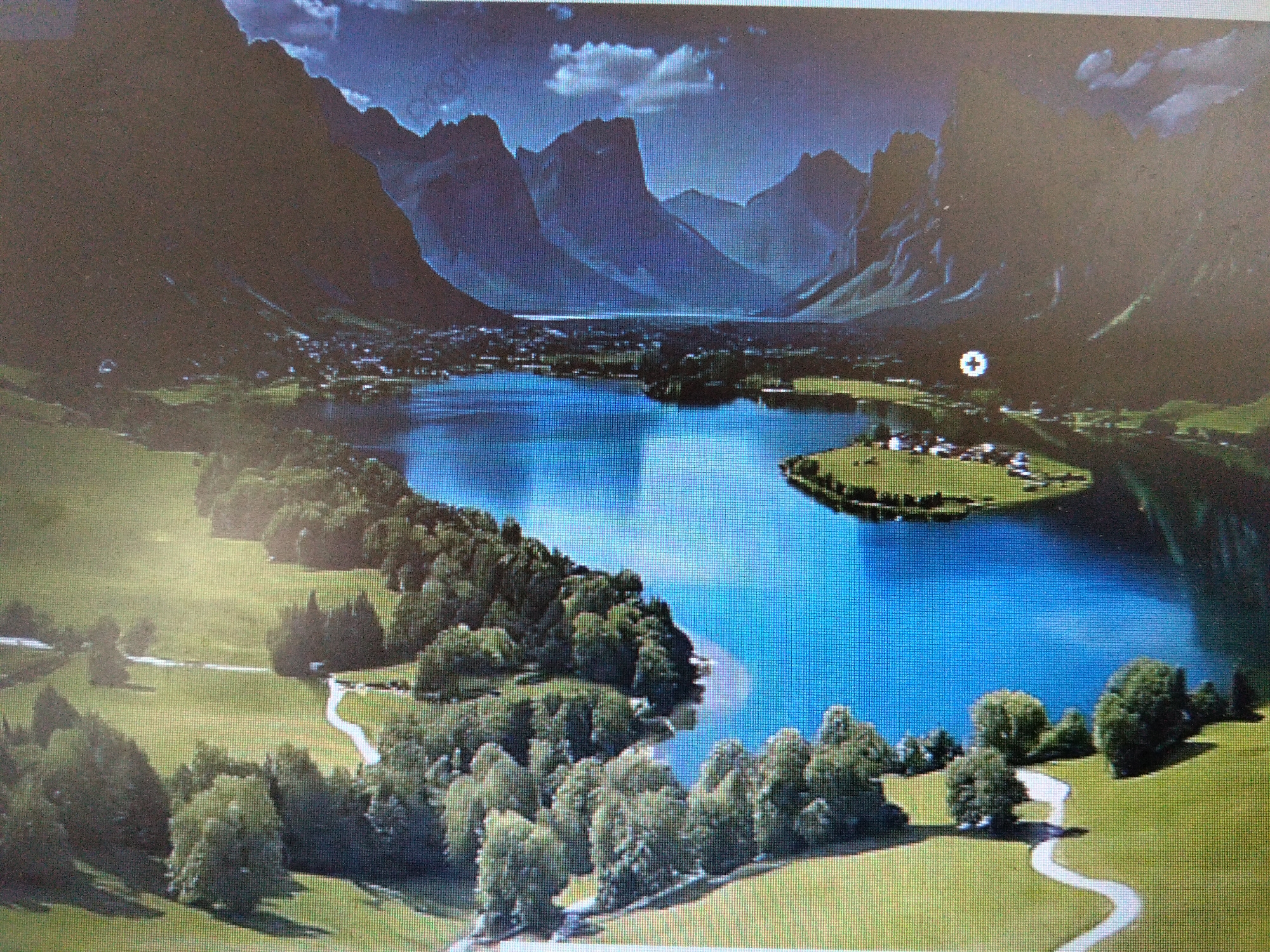This is an aerial photograph of a picturesque mountain landscape with a clear blue lake. The lake is surrounded by lush green trees and rolling hills. In the distance, majestic mountains rise, their peaks partially obscured by dramatic clouds. A small village with white-roofed buildings is nestled on the shore of the lake, and a few scattered houses are visible on a small island in the center of the water. Winding white paths traverse the grassy areas, suggesting hiking trails. The image does not depict the city of Cotgadong, Indonesia, or any elements typically associated with an Indonesian urban environment.