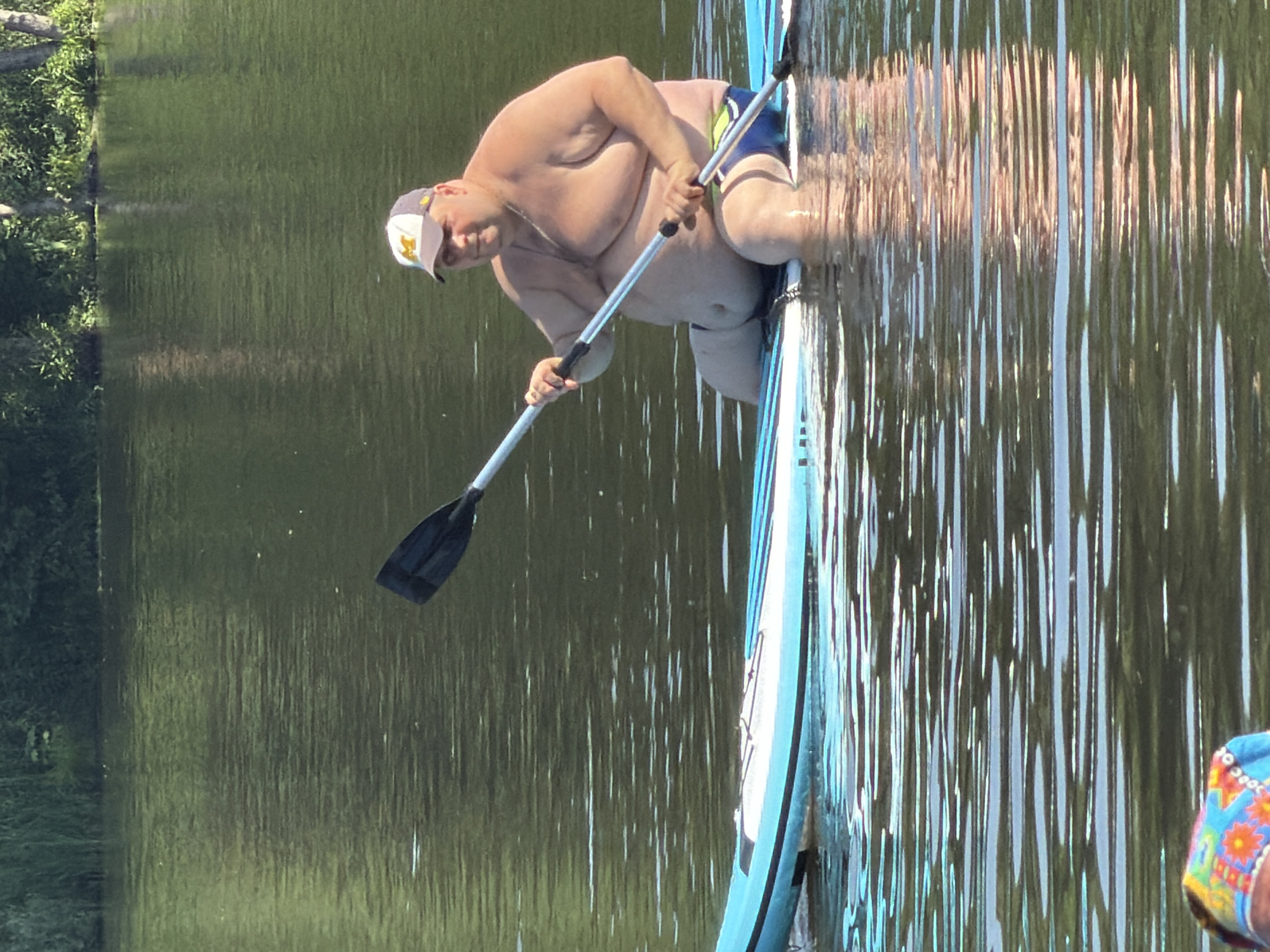 An overweight man wearing a baseball cap and swim trunks sits on a stand-up paddleboard on a body of water. He is holding a paddle, with the blade extended to the left of the frame. The water is calm with reflections of the surrounding greenery. The man is looking towards the camera. In the foreground, a colorful patterned fabric is visible. The background shows a line of trees.