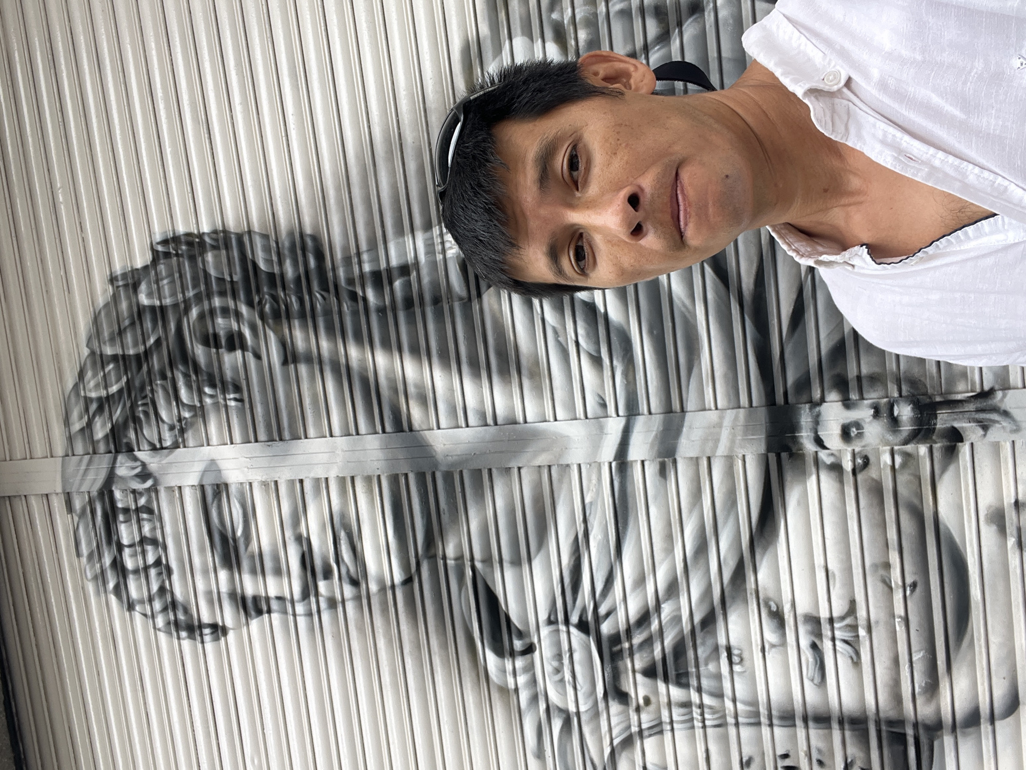 An adult Asian male is taking a selfie in front of a mural painted on a metal roll-up door. The mural is a black and white depiction of a classical statue's face and upper body. The man is wearing a white short-sleeved collared shirt, sunglasses pushed up onto his forehead, and a black headband or earbud cord visible behind his right ear. His expression is neutral, and he is looking directly at the camera. The metallic slats of the roll-up door are clearly visible through the mural and behind the man, creating a textured background. The scene appears to be outdoors, possibly in an urban setting, given the street art and the nature of the shuttered storefront. While the specific location in Tarragona, Spain is not explicitly visible, the style of street art and architecture might be characteristic of certain areas in the city.