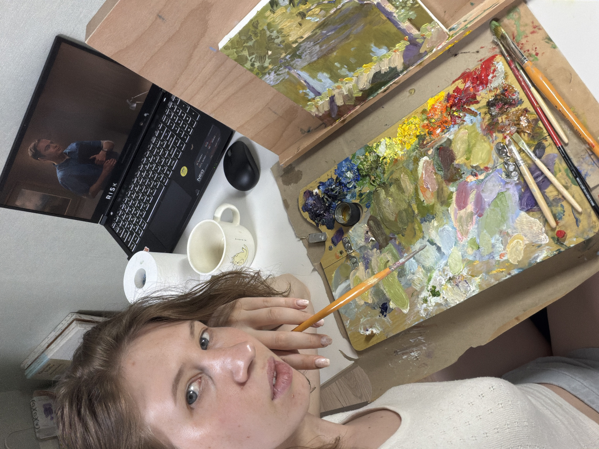 The image shows a young woman with brown hair and blue eyes, looking at the camera while resting her head on her hand. She is wearing a white top. In front of her is an artist's palette with a variety of colorful paints, and several paintbrushes. To her right, a laptop is open, displaying a video of a man in a blue shirt. To her left, a roll of toilet paper and a white mug with a yellow cat design are visible. In the background, there are some books and what appears to be an easel with a partially completed painting of a natural scene. The setting is likely an indoor art studio or workspace. There is no information in the image that suggests the location is in Dongducheon-si, South Korea.