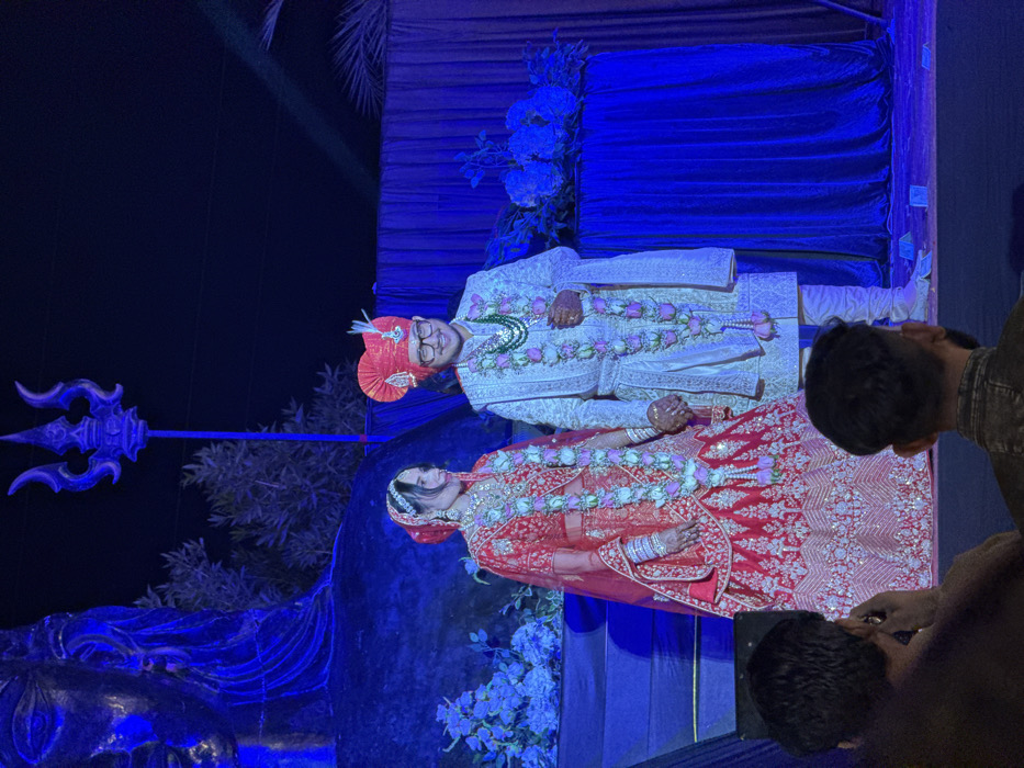 The image captures an elaborately decorated scene, likely an Indian wedding or engagement ceremony, featuring a couple in traditional attire. The photograph is oriented vertically, with the subjects appearing to be standing upright against a dark, blue-lit background.

In the center of the frame are the bride and groom. The groom, positioned on the left, is wearing a white or cream-colored sherwani with intricate gold embroidery and embellishments, possibly sequined or beaded. He has a bright red turban (pagdi) adorned with a decorative piece and is wearing glasses. Around his neck are multiple floral garlands, predominantly white and light pink roses. He is smiling and appears happy.

The bride, standing to the right of the groom, is dressed in a vibrant red lehenga (skirt) and choli (blouse) set, heavily embroidered with gold and possibly silver threads, creating a rich, shimmering texture. Her head is covered with a matching red dupatta, and she wears elaborate jewelry including a necklace, earrings, a maang tikka on her forehead, and numerous bangles on her wrists. Her hands are adorned with mehndi (henna). She also wears multiple floral garlands, similar to the groom's, around her neck. Her expression is less clear, as she appears to be looking slightly downwards or to the side.

The couple is standing on what appears to be a raised platform or stage. The backdrop behind them consists of dark blue draped fabric, possibly velvet, which is strongly illuminated with blue lighting, giving the scene a cool, dramatic glow. Artificial floral arrangements, resembling blue hydrangeas, are visible on the backdrop.

To the left of the couple, a prominent, large, stylized trident (trishul) stands upright, also bathed in blue light. This symbol suggests a connection to Hindu mythology, particularly Lord Shiva. Below the trident, there's a dark, sculpted element, possibly part of a larger statue or stage decoration, also illuminated in blue.

On the right side and in the foreground, several other individuals are partially visible, suggesting they are observers or attendees. Their heads and shoulders are cropped, and they appear to be looking towards the couple. One person in the bottom right corner is holding a mobile phone, likely capturing photos or videos of the event. The overall lighting is dim, emphasizing the blue hues and spotlighting the couple. The setting suggests an evening event with carefully orchestrated lighting and decor.