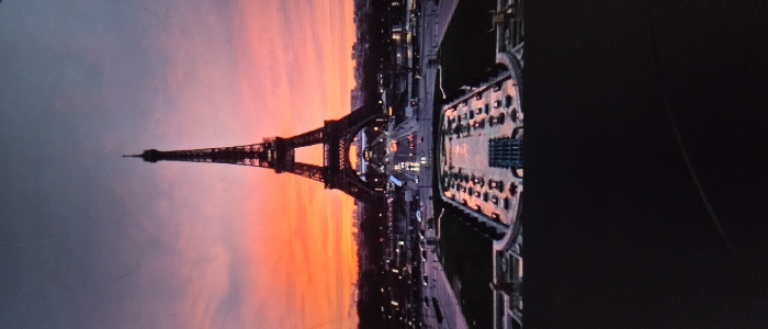 The image displays a prominent, dark-colored lattice tower positioned centrally against a vibrant sunset or sunrise sky. The sky exhibits a gradient of intense orange, red, and pink hues at the horizon, transitioning to a darker blue-grey above. In the midground, a multi-lane urban thoroughfare is visible with active vehicle traffic, indicated by blurred streaks of headlights and taillights. Various city buildings, some illuminated, line the street and extend into the distant background. The immediate foreground features a series of structured, multi-level water basins and surrounding landscaping, reflecting the colors of the sky and the city lights. No individual persons are distinctly discernible. This depicted scene is located in Giengen an der Brenz, Germany.