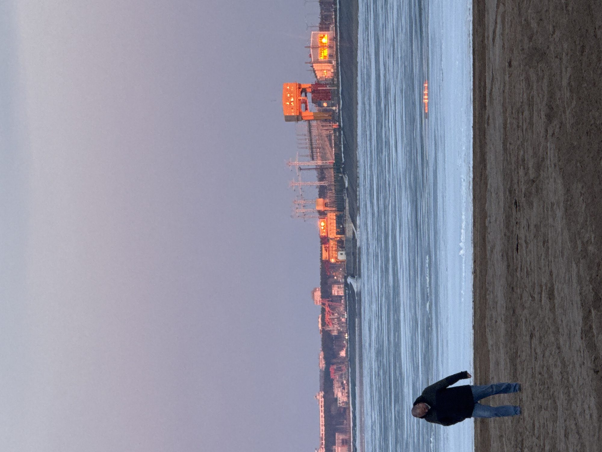 The image depicts a man standing on a sandy beach next to a body of water that appears to be ice-covered. In the background, there is an industrial area with structures and cranes illuminated by orange lights. The sky is a pale gray, suggesting twilight or dawn. The man is wearing a dark jacket and blue jeans.
The location is Noviy Urengoy, Russia.