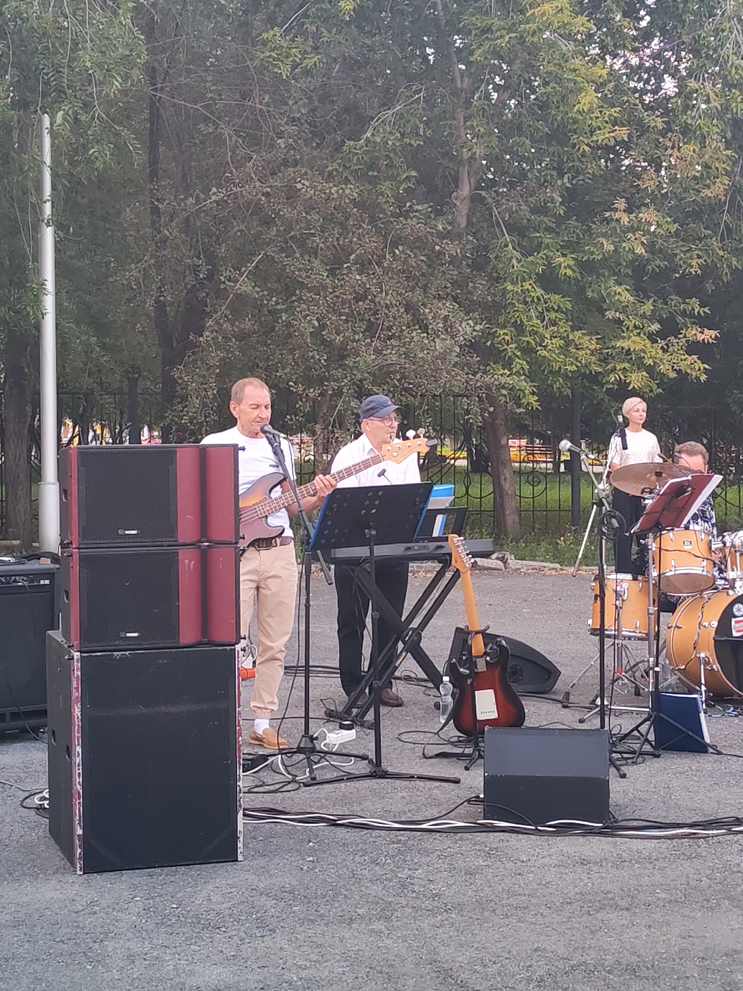 A group of four musicians perform outdoors in front of a backdrop of trees. From left to right, a man in a white t-shirt and tan pants plays a bass guitar. To his right, a man in a white collared shirt, dark pants, and a blue baseball cap plays a keyboard. To his right, a woman with short blonde hair in a sparkly white top stands and sings into a microphone. To her right, a man in a patterned shirt and glasses plays a drum set. In the foreground, large black speakers are stacked on the left side of the image. A keyboard stand and an electric guitar are visible in the center. Cables snake across the asphalt ground. The scene appears to be set in a park or public space.
