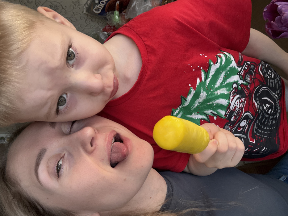A close-up photograph depicts an adult female and a young male child in an indoor residential setting in Chaikovsky, Russia. The female, positioned on the left, has light-colored hair and wears a dark grey top; her left eye is closed, and her tongue is extended in a playful gesture. The male child, positioned on the right, has light blonde hair and green-grey eyes. He wears a red t-shirt featuring a green Christmas tree with white accents and a graphic of Santa Claus driving a red monster truck. The child holds a bright yellow, cylindrical popsicle in his right hand. His expression is directed towards the camera, appearing neutral to slightly somber. The blurred background reveals a light-colored, possibly patterned wall, a dark package with "Choco" visible text, and the partial view of a purple tulip in the lower right corner.
