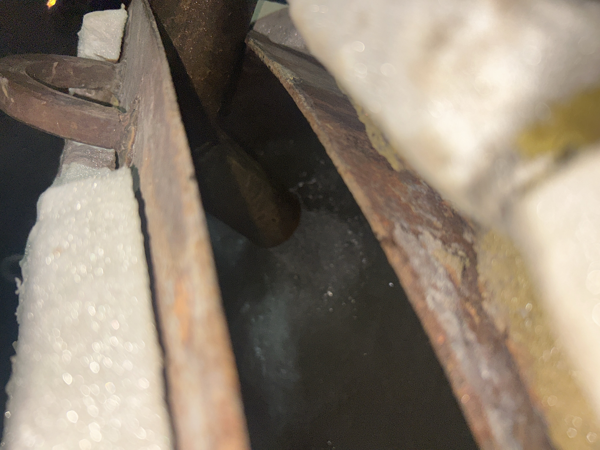 The image depicts a close-up, low-angle view of what appears to be a metal object, possibly a part of industrial machinery or a structural component, showing signs of rust and wear. The metal is thick and has a dark, weathered surface. It is partially covered by what looks like insulation material, which is white and foamy. In the background, there is a dark, possibly liquid-filled trough or channel, with some particulate matter visible on its surface. The lighting is dim, with some specular highlights suggesting a light source from above and to the side. The overall impression is one of decay and disuse. There are no people or discernible activity. The image does not contain any elements that would allow for identification of its specific location within Krasnodar, Russia, or any city.