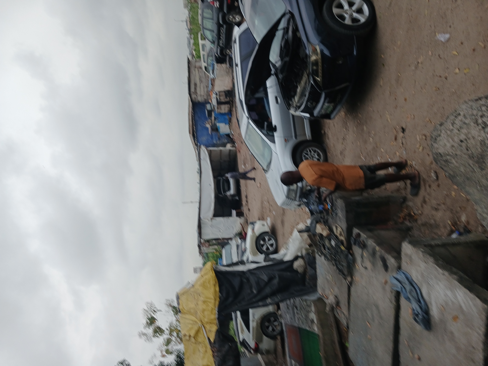 The image depicts an outdoor scene in what appears to be a cluttered area with several parked cars and makeshift structures. In the foreground, a man wearing an orange shirt and dark shorts stands near a dark-colored car with its hood open. The car appears to be undergoing some form of repair or inspection. Behind him, a silver car is parked, also with its hood up. Further in the background, more cars are visible, along with several sheds and tarpaulin structures, suggesting a junkyard, a car repair lot, or a similar informal commercial or industrial area. The ground is rough and sandy, with debris scattered around. The sky is overcast and cloudy, indicating a dull or potentially rainy day. The overall impression is one of a busy, somewhat unkempt, and functional environment.