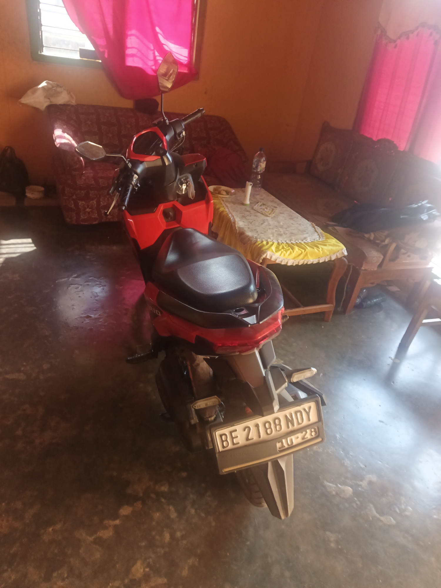 The image shows the rear view of a red and black scooter parked indoors. The scooter has a license plate that reads "BE 2188 NDY 10-28". In the background, there is a patterned couch and a table covered with a yellow cloth. The room has orange walls and bright pink curtains covering a window. The floor appears to be concrete. This scene is likely located in a residential or storage area within Terbanggi Marga, Indonesia.