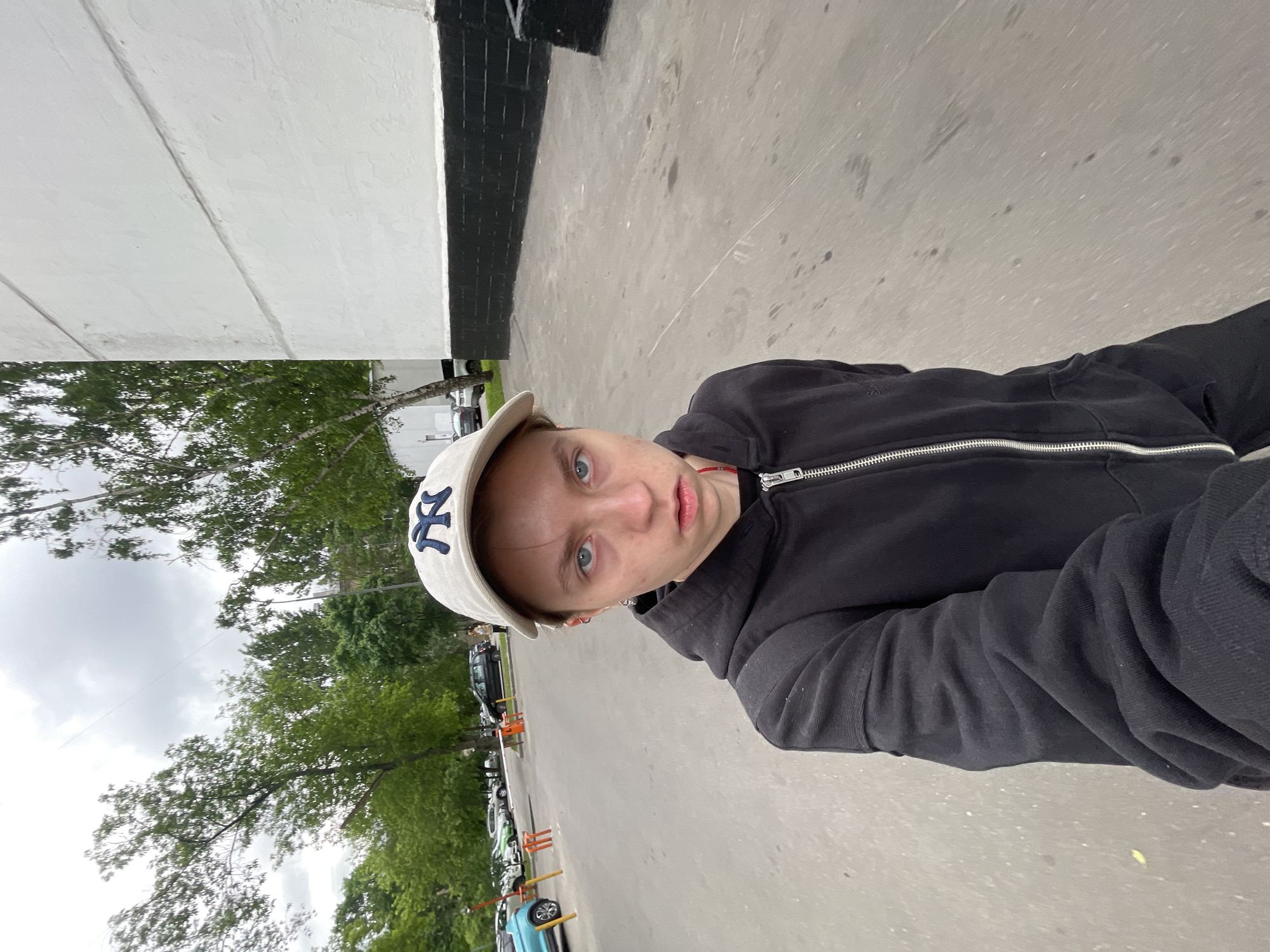 A person is standing in an outdoor parking area or street, likely in Moscow, Russia. They are wearing a black zip-up hoodie and a light-colored baseball cap with a "NY" logo. The person is looking at the camera. The sky is overcast. In the background, there are trees and parked cars. A building is visible on the left side of the image.