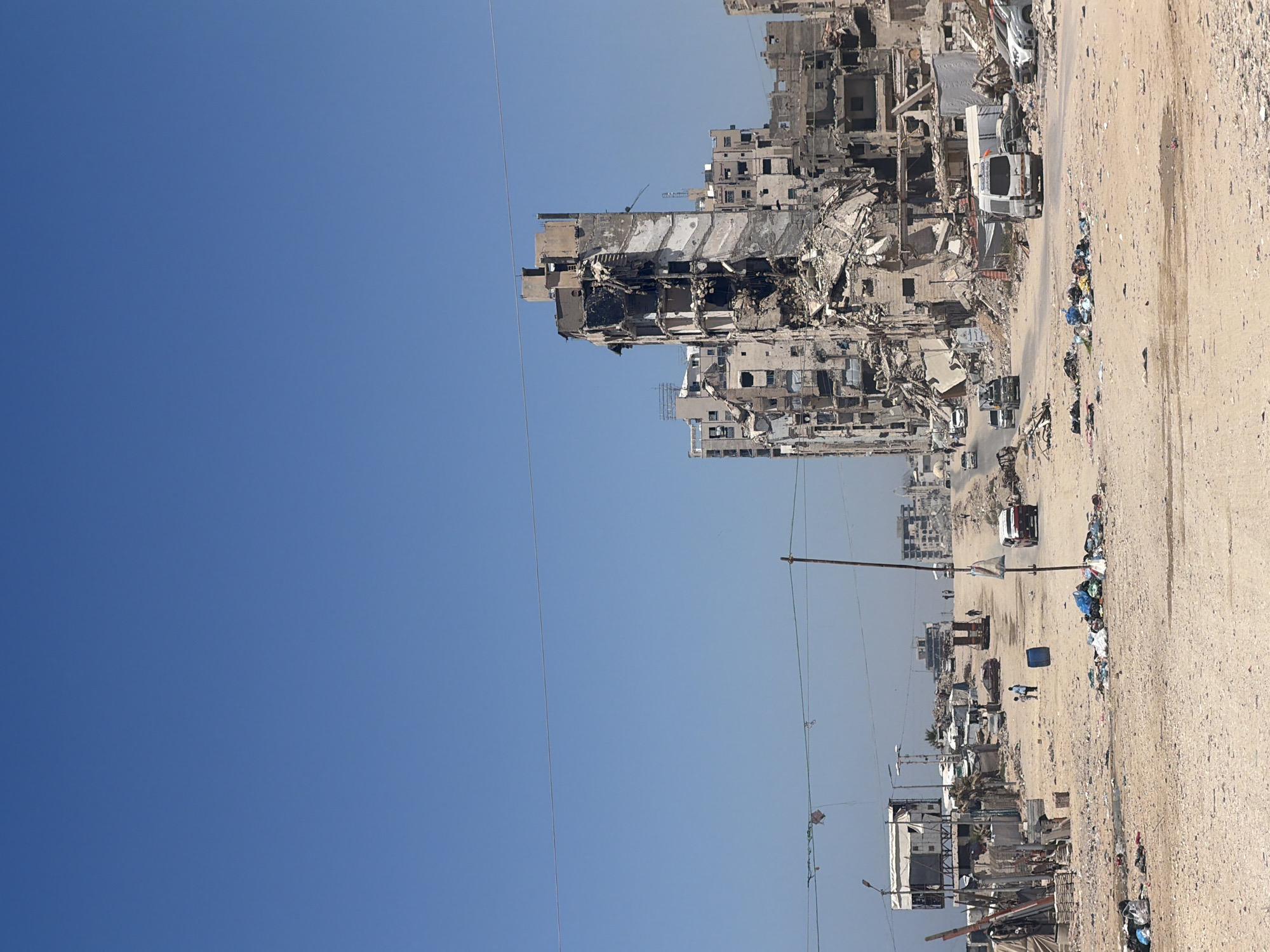 This is a wide shot of a devastated urban environment under a clear blue sky. The foreground is a dirt and gravel road littered with debris, including scattered trash bags and plastic sheeting. In the middle ground, several cars are visible, including a white van with Arabic writing on its side, and a red car. People are walking along the road and near some temporary structures that appear to be makeshift shelters.
The background is dominated by heavily damaged multi-story buildings. The building on the right is severely damaged, with large sections of its facade missing and debris scattered around its base. Other buildings in the background also show significant structural damage. The overall impression is one of destruction and disarray.