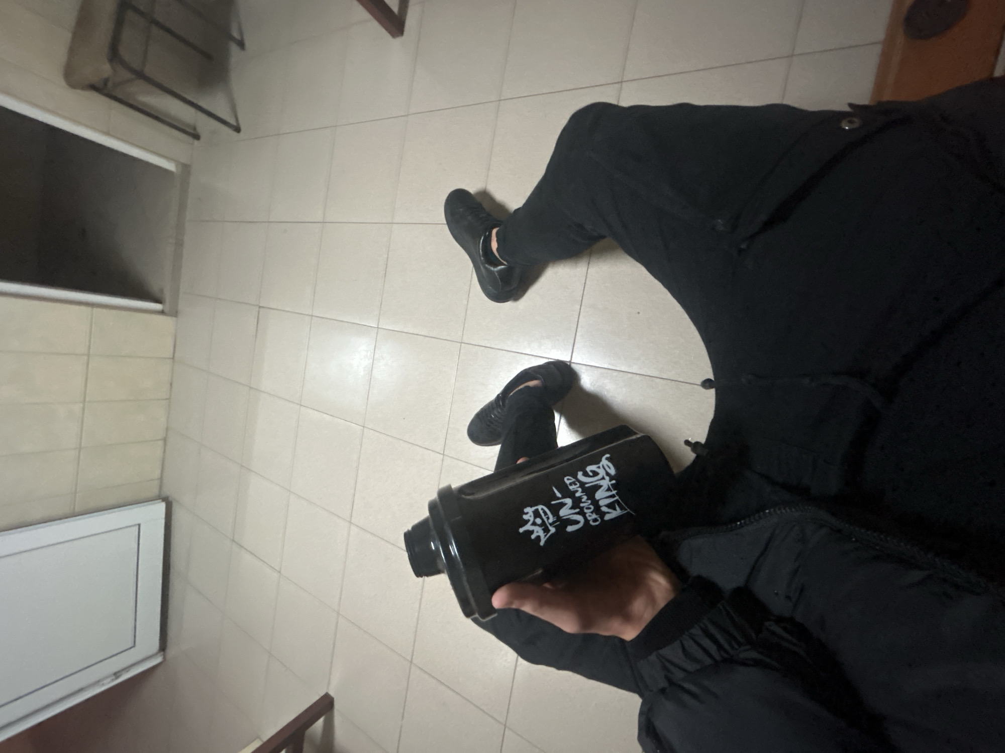 The image shows a person sitting on a tiled floor. The person is wearing black pants, black shoes, and a black jacket. They are holding a black container with white writing on it, which includes the words "UN-CROWNED KING". In the background, there is a window and some furniture. The location appears to be indoors. The location of the image is Zlatograd, Bulgaria, as this information was provided.