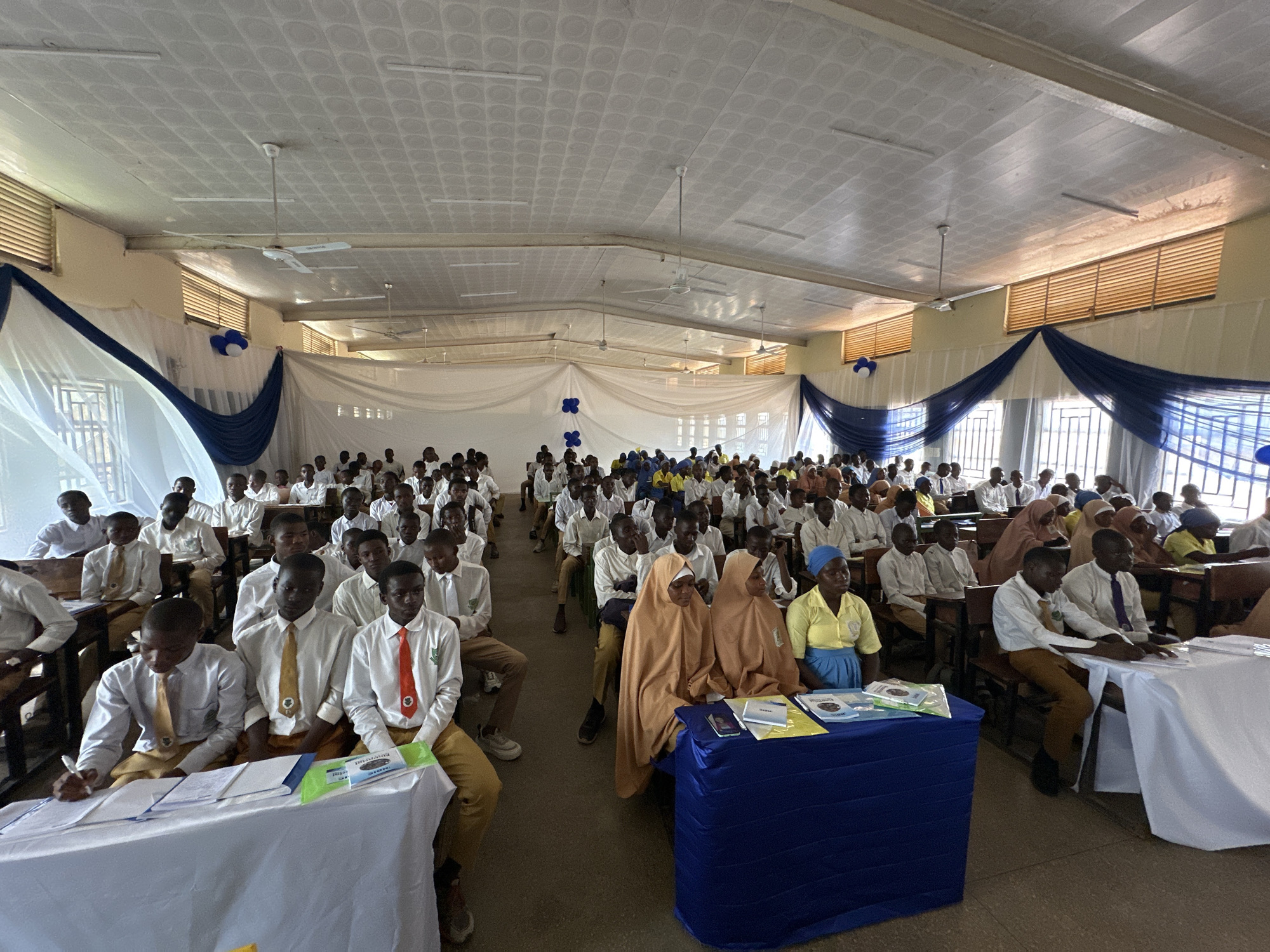 This image captures a large indoor gathering, likely an academic or educational event, in what appears to be a school hall or assembly room. Numerous young people, predominantly male students, are seated in neat rows at wooden desks. They are dressed in white long-sleeved shirts, with many wearing ties of various colors such as yellow, orange, red, and purple, paired with light brown or khaki trousers, consistent with school uniforms.
At the front of the room, on the left, a student is actively writing on papers placed on a white-covered table, which also holds green folders and books. On the right side, two women in light brown hijabs and long garments, alongside another woman in a yellow top, blue skirt, and blue head covering, are seated behind a table draped in blue fabric. This table also displays books and educational materials; visible text on one item includes "FINANCIAL" and "topic," with another book partially showing "Pillars of...".
The room is decorated with white sheer fabric draped artistically across the upper sections of the walls and windows, accentuated with swathes of dark blue fabric. Blue and white balloons, and blue fabric cut into star or cross shapes, are used as additional embellishments. The ceiling features a patterned white paneling, multiple ceiling fans (one clearly marked "ORIENTAL"), and fluorescent lighting. Natural light streams in through high windows equipped with horizontal blinds and protective metal bars, indicating a daytime event. The overall scene suggests an organized and attentive gathering, possibly a seminar, workshop, or competition.