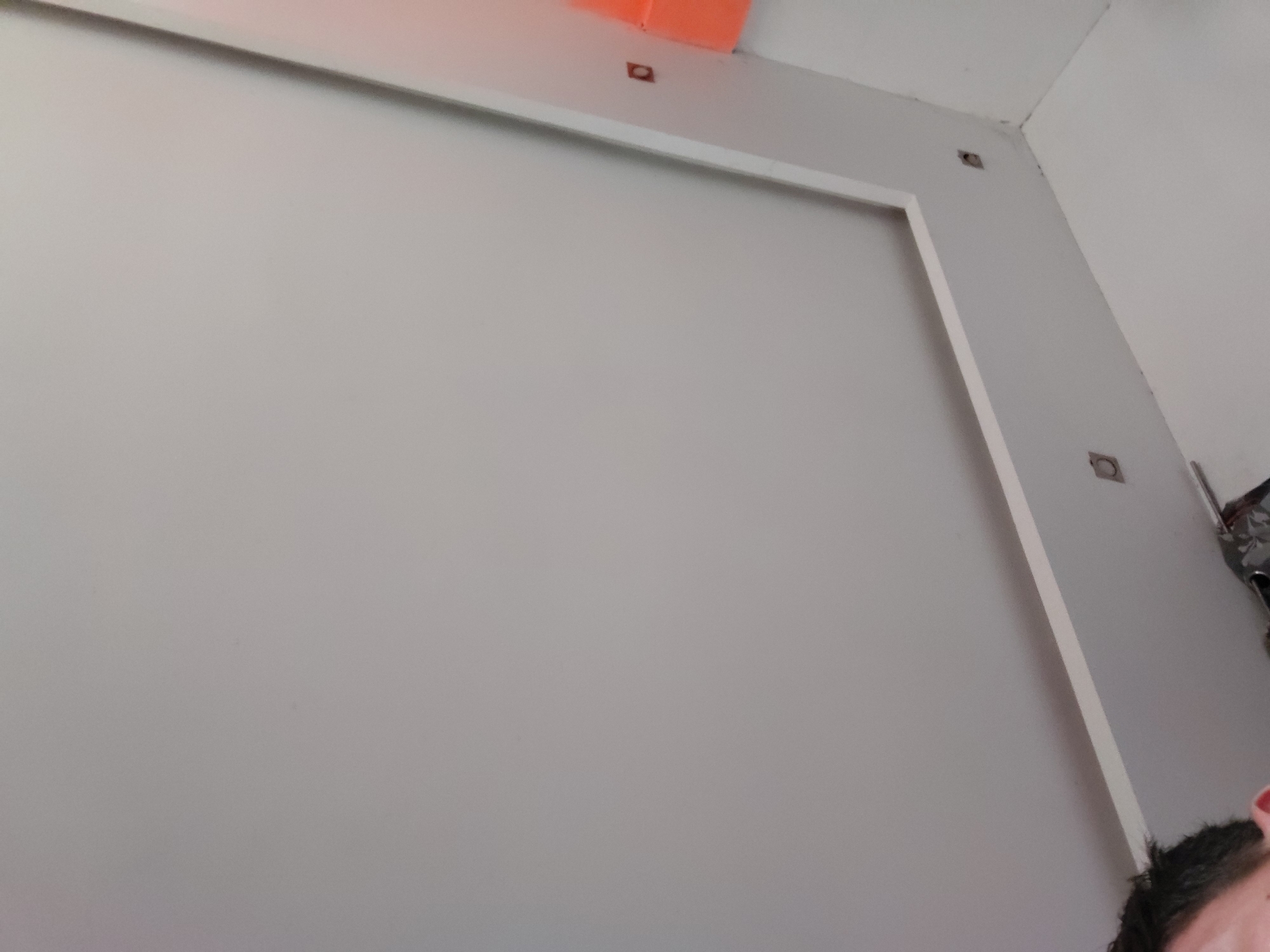 This image depicts an interior scene, likely a room undergoing construction or renovation, located in Santa Ana, Venezuela. The central focus is a large, plain light gray wall adorned with a prominent white trim that outlines a rectangular section, creating a framed panel effect.
Several small, rectangular cutouts or holes are visible on the gray wall and the adjacent white ceiling/upper wall. These appear to be prepared openings for electrical outlets, switches, or light fixtures; some show dark recesses, while one in the upper left contains a small, reddish-orange component.
In the upper-left corner of the frame, a bright orange object or surface is partially visible, possibly a section of the ceiling or an adjacent structural element. The white ceiling or upper wall area displays minor imperfections, particularly near the corner joint with the gray wall. On the far right side, near the top, a small portion of a metallic object with a patterned design (possibly floral) can be seen.
A person's head, showing dark hair, an ear, and a red fingernail, is partially visible in the bottom-right corner, indicating the presence of an individual, likely the photographer. The lighting is bright and uniform, suggesting an indoor setting, though the specific time of day is not discernible. No text is visible within the image. The overall impression is that of a functional space in the process of being fitted out or upgraded.