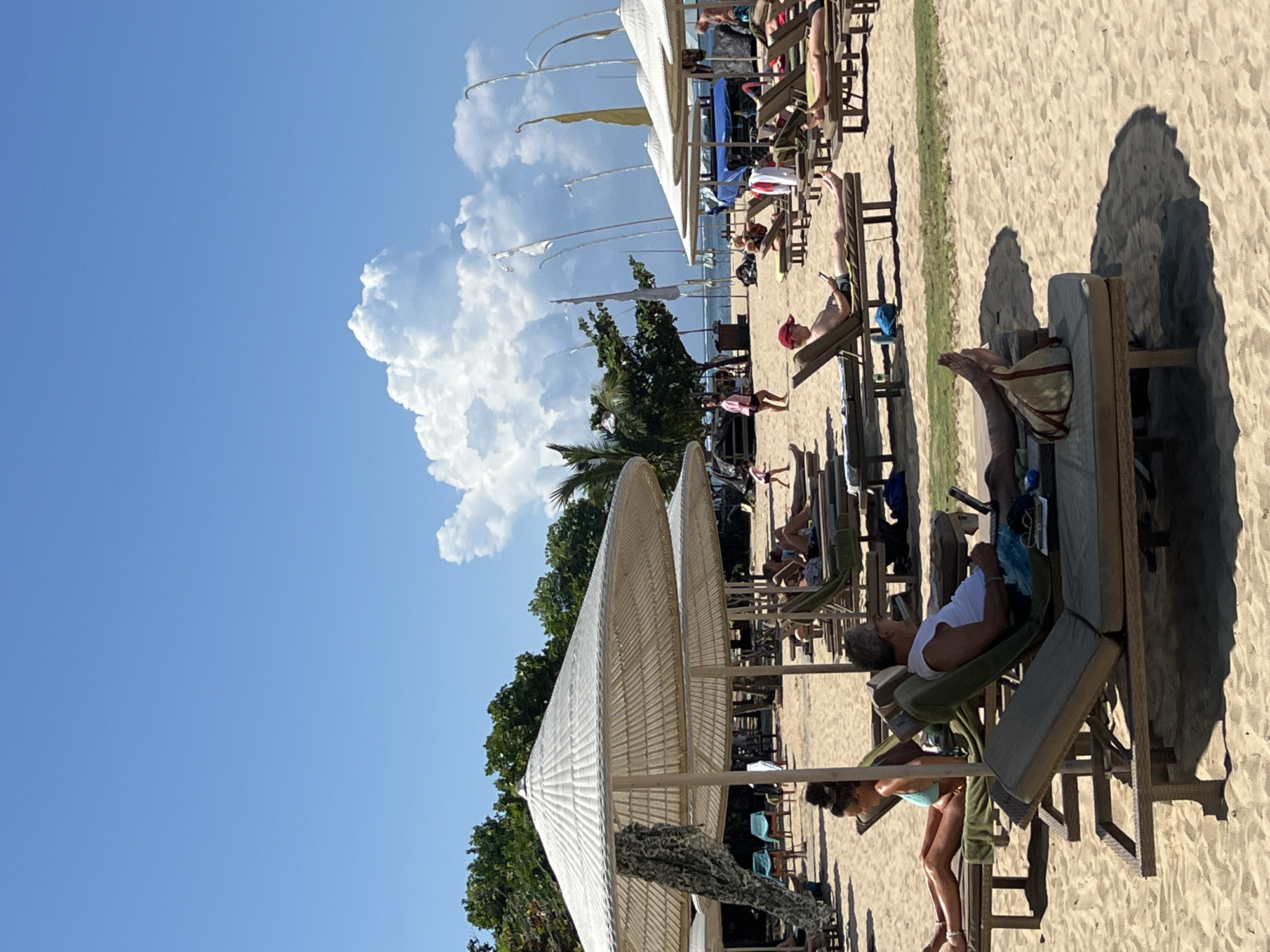 The image depicts a beach scene in Denpasar, Indonesia, likely in the Seminyak or Kuta area, known for its resorts and sandy shores. The foreground is dominated by rows of empty and occupied lounge chairs on a light brown sand beach. Several large, off-white umbrella shades are set up, providing cover for some of the chairs.
In the middle ground, several people are seen relaxing on lounge chairs. One man in a red cap is looking at his phone. Another man with gray hair and a white shirt is using a laptop. A woman in a turquoise bikini is reclining on a chair further back. Other individuals are visible in lounge chairs or walking on the sand.
The background features more lounge chairs, some umbrellas, and palm trees. In the distance, the ocean is visible, with several white flags on poles lining the shore. The sky is a clear blue with a large, fluffy white cloud formation. A small child is running in the sand in the middle distance. The overall atmosphere suggests a sunny day at a popular beach resort.