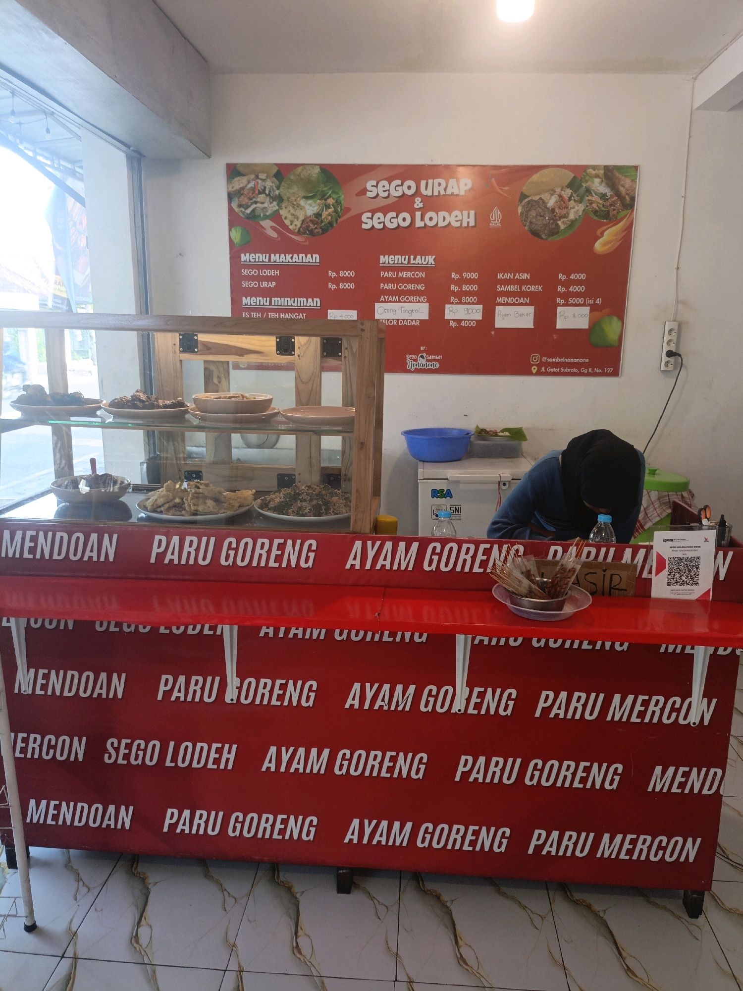 A street food stall named "Sego Urap & Sego Lodeh" is depicted in a medium shot, positioned at Jl. Gatot Subroto, Gg II, No. 127 in Probolinggo, Indonesia. The stall has a red banner at the front displaying menu items like "Mendoan," "Paru Goreng," and "Ayam Goreng" in white lettering, repeated multiple times. Behind the counter, food items are displayed in a glass-fronted cabinet. A woman wearing a black hijab and blue long-sleeved shirt is partially visible behind the counter, interacting with the food. The stall is equipped with a cooler, a blue plastic basin, and a QR code for payment. The overall scene suggests a busy, casual dining environment.