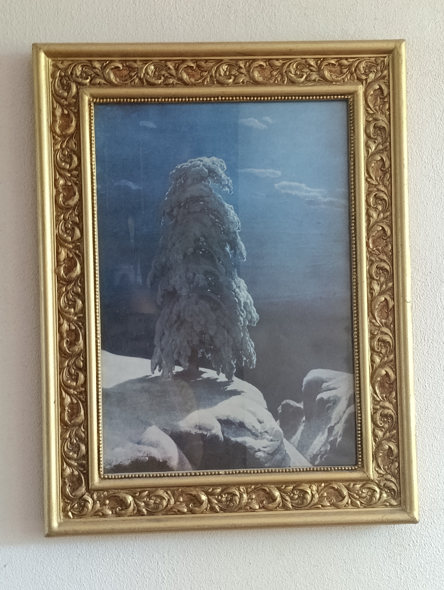 A framed painting of a snow-covered tree on a rocky outcrop under a blue sky with clouds is displayed on a white, textured wall. The painting is rectangular and oriented vertically. The frame is ornate, gold-colored, and appears to be made of wood. The painting depicts a lone, dense evergreen tree, heavily laden with snow, dominating the center of the composition. The background features a gradient of blue, suggesting a winter day with a bright sky. In the foreground, snow-covered rocks and snowdrifts create a rugged landscape. The overall scene is one of a serene, wintry natural setting. The image is taken from a frontal, slightly angled perspective, showing the painting hanging on the wall.