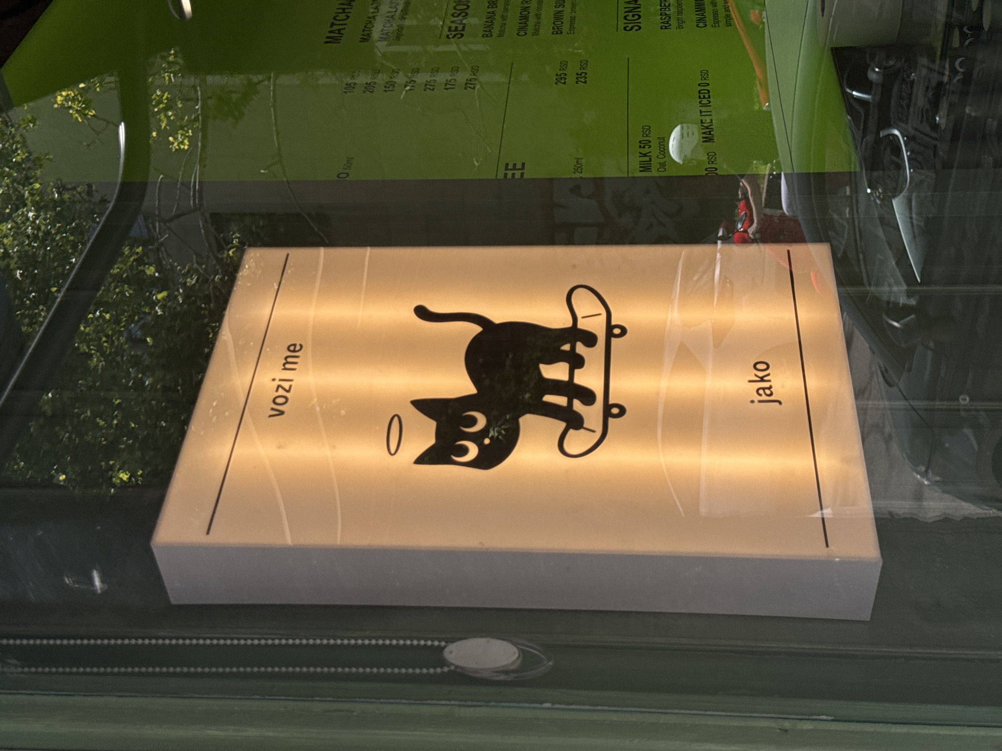 The image captures a storefront window in Belgrade, Serbia, reflecting the surrounding environment and displaying interior signage. A prominent backlit sign features a stylized black cat wearing a halo, standing on a skateboard. The text "vozi me" and "jako" are visible on the sign. To the right, a menu board is visible through the window, with text including "matcha", "seasonal", "milk", and "signa". Reflections in the glass show trees with green foliage and sunlight, as well as a green vehicle and car interiors. The exterior of the building appears to be painted green. The overall impression is of a bright, modern cafe or shop.