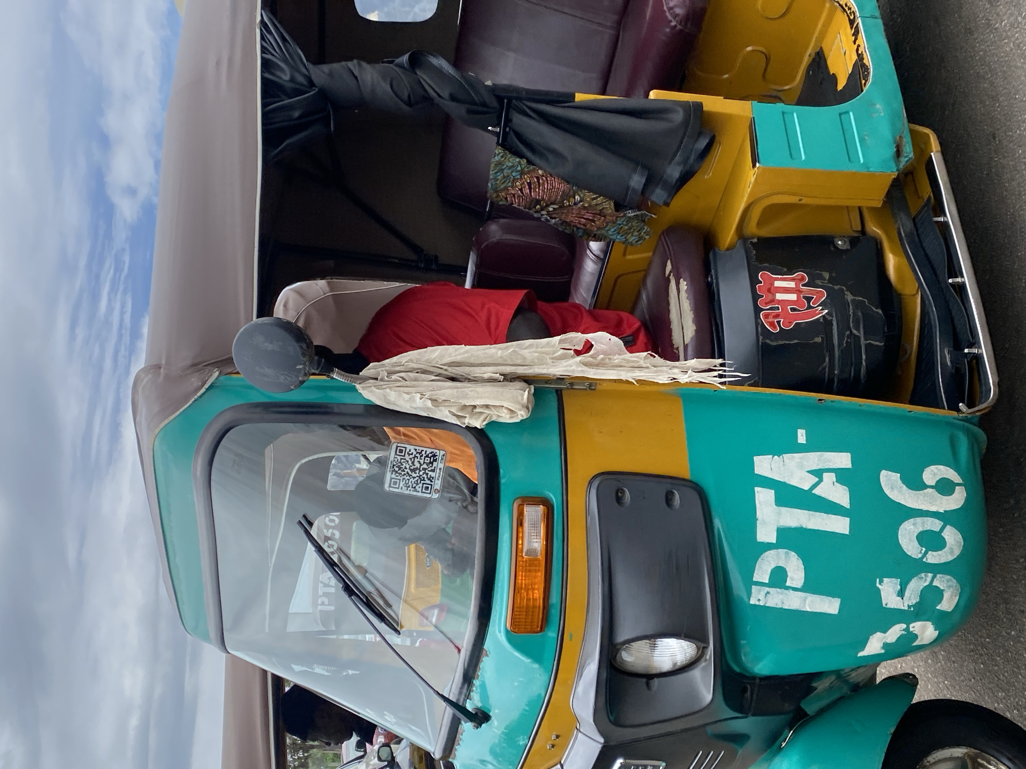 In Rafin Par, Nigeria, a teal and yellow three-wheeled auto-rickshaw, identified with the stencil markings "PTA-3506" on its side, is parked on an asphalt road under a cloudy sky. The vehicle's interior, visible through the windshield, contains a driver wearing a black beanie and a red shirt. A QR code is affixed to the windshield, and a piece of torn off white cloth hangs from the side mirror. The auto-rickshaw has maroon-colored seating and a brown fabric roof. A black spare tire is visible inside the passenger compartment.