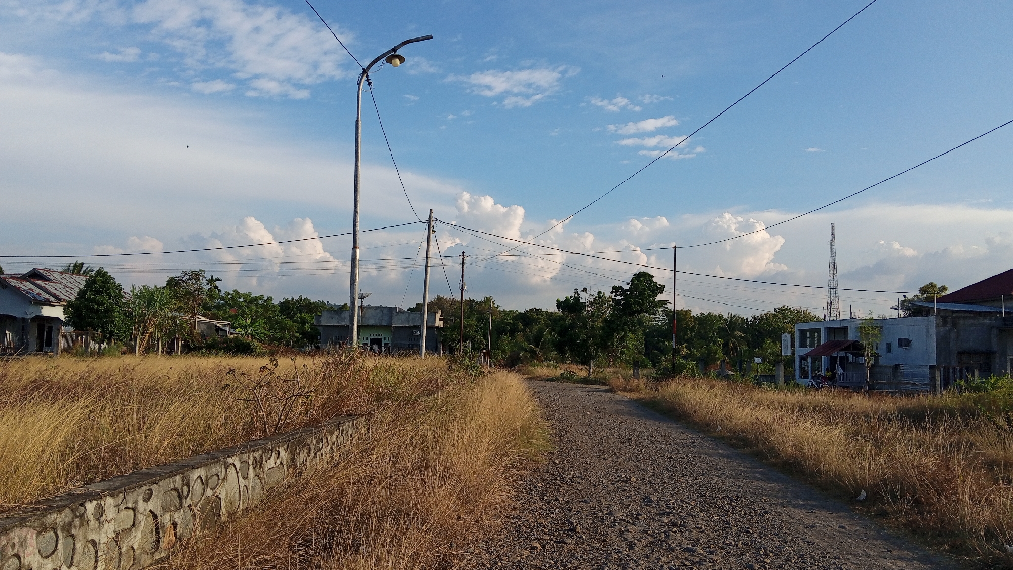 This is a photograph of a dirt road in Geulanggang Baro, Indonesia, bordered by dry, golden grass and a low stone wall on the left. The road curves gently into the distance, with a few scattered rocks visible on its surface. On the left side of the road, there is a house with a corrugated metal roof and what appears to be an unfinished concrete facade. Further back, amidst lush green trees, more houses are visible, one with a light green exterior and a satellite dish.
To the right of the road, the dry grass continues, with a modern-looking building situated behind it. This building has white walls, a red-tiled roof, and several windows. A tall communication tower stands in the background, behind several trees. Overhead, a bright blue sky is dotted with large, fluffy white clouds. A street lamp stands on a tall pole on the left side of the road, and power lines crisscross the sky, supported by utility poles. The overall impression is of a rural or semi-urban landscape bathed in the warm light of what appears to be late afternoon. No people are clearly discernible in the image, though there might be some figures in the shade of the building on the right.