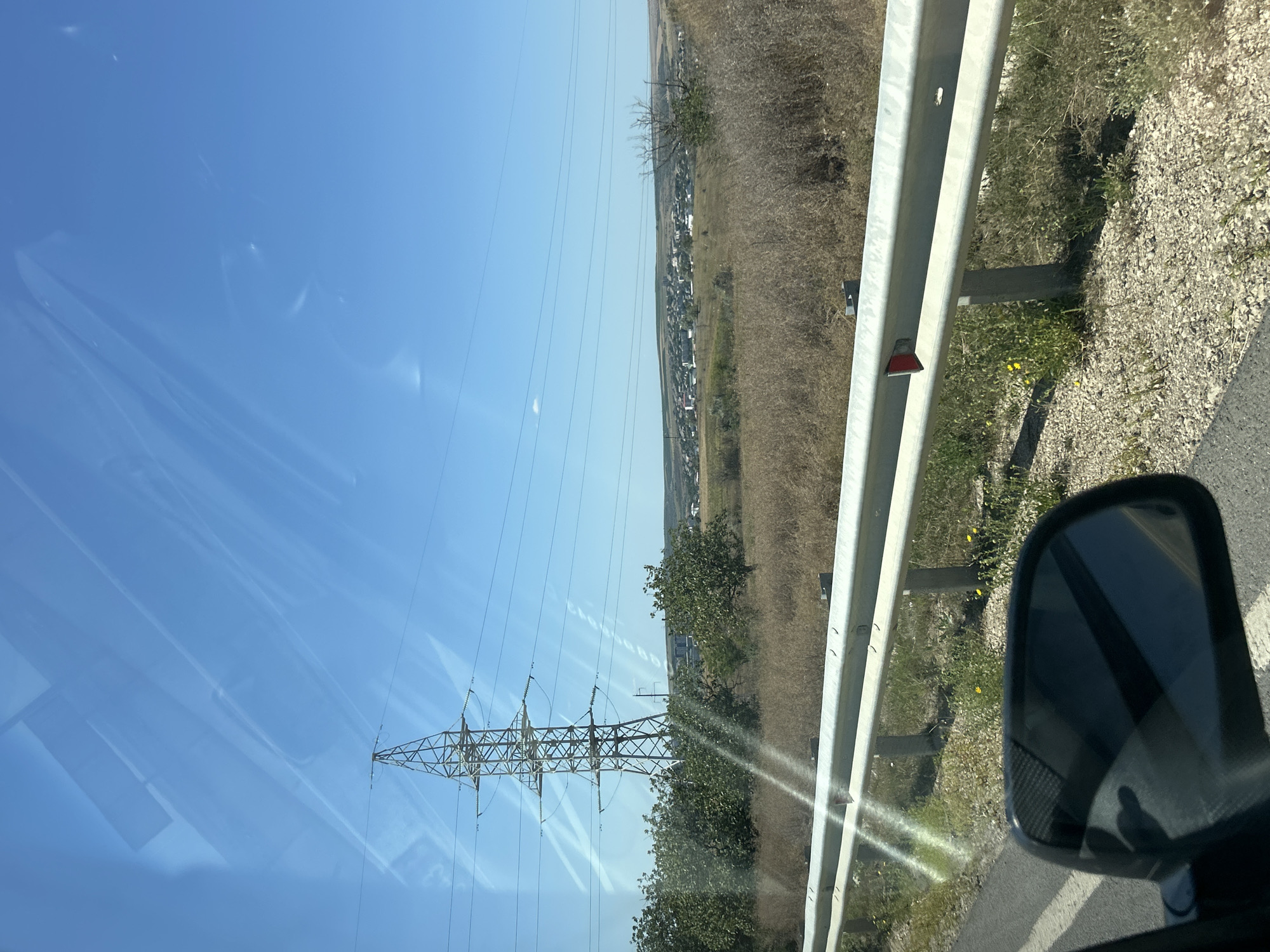 An eye-level, medium shot captures a rural landscape under a clear blue sky, likely on the outskirts of Comrat, Moldova, given the context. In the foreground, a portion of an asphalt road is visible on the left, with white dividing lines. A metallic guardrail runs horizontally across the middle of the frame, with its reflection visible in the side-view mirror of what appears to be a vehicle, also on the left. Beyond the guardrail, a dry, grassy field stretches out, dotted with sparse green bushes and trees. In the middle ground, a prominent high-voltage electricity pylon stands tall, with its power lines extending across the sky. Further in the distance, a small town with white buildings and rooftops is nestled among rolling hills. The overall scene depicts a sunny day in a semi-arid, undeveloped area, characterized by infrastructure and a small settlement.