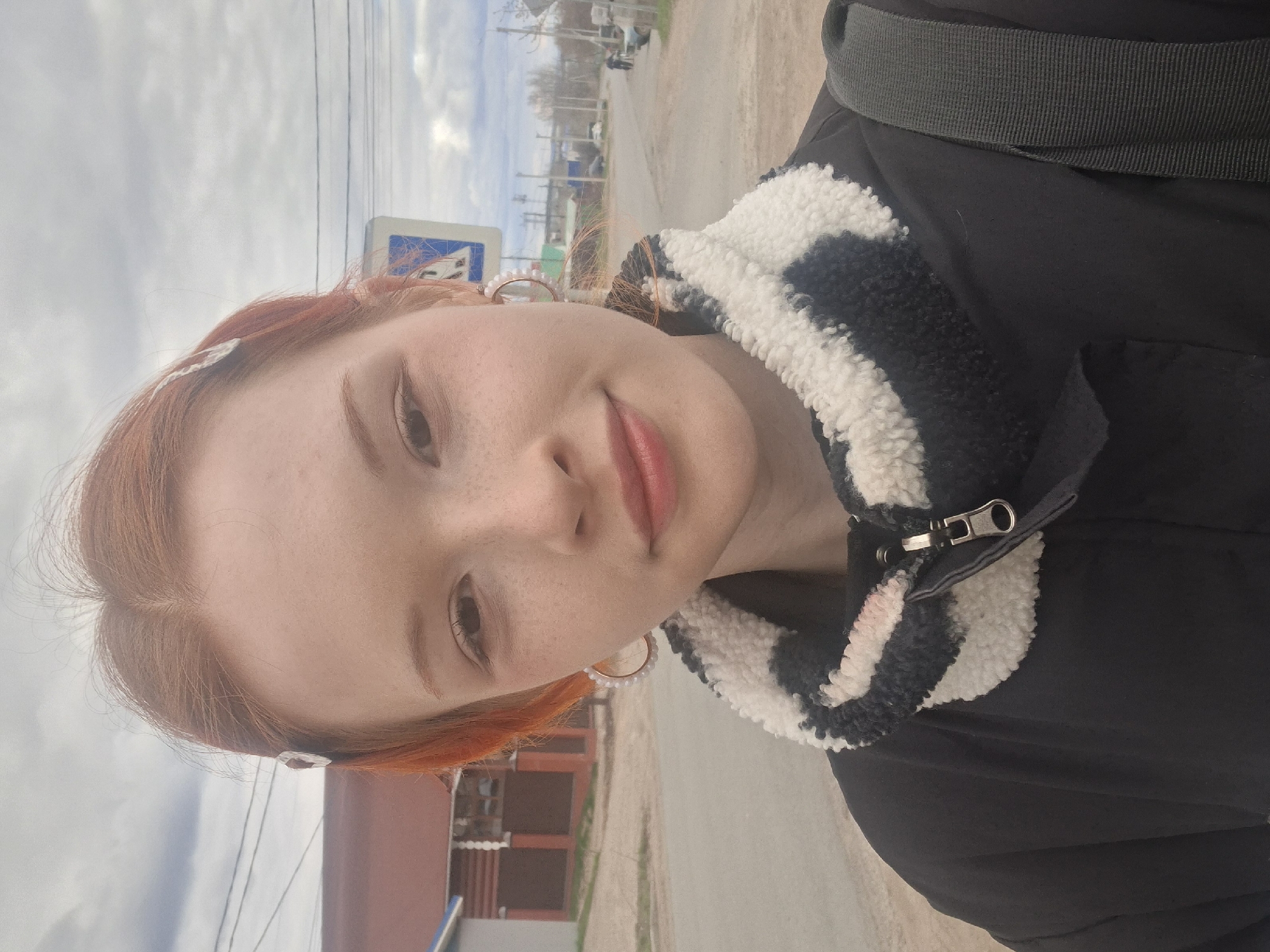 An outdoor portrait shot in Chaikovsky, Russia, features a young woman with reddish-orange hair and fair skin, lightly dusted with freckles, posing with a soft smile. She wears a black jacket with a distinct black and white striped, fuzzy or sherpa-like collar. Her lips are tinted with a reddish-pink lipstick, and she has visible pearl-edged hoop earrings and a small, clear or white decorative hair clip. A dark grey strap, possibly from a bag, is visible over her shoulder.
The background, partially out of focus, suggests a roadside or street setting on an overcast day. Visible elements include a light grey, cloudy sky filling the upper portion of the frame. In the mid-ground, utility poles with wires stretch across the view, and a partially obscured blue and white traffic sign is visible. Distant buildings, one appearing reddish-brown, line what seems to be a road or path. Bare trees or shrubs indicate either late autumn, winter, or early spring. The overall lighting is soft and diffuse, characteristic of an overcast sky. No discernible text is visible in the image.