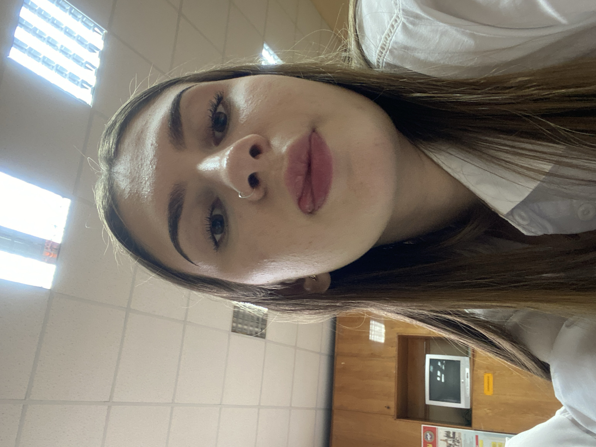 The image shows a close-up of a young woman's face, with her tongue sticking out slightly. She has dark brown hair and is wearing a white shirt. The background appears to be an indoor environment with a tiled ceiling and fluorescent lighting. There are wooden cabinets and a monitor visible in the background. The woman is wearing a nose ring. There is no indication of the location being the city of Дьяково, Russia.