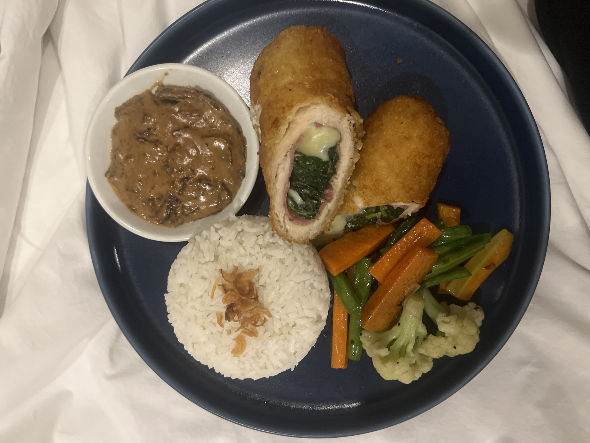 A close-up, overhead shot captures a complete meal artfully arranged on a dark blue, circular plate, set against a bright white fabric background, likely a bedsheet or tablecloth, suggesting a casual indoor dining setting such as a hotel room or home in Kuta Selatan, Indonesia.
The central focus of the meal is two pieces of golden-brown, breaded and fried chicken. One piece is halved, revealing its savory filling: a layer of tender chicken wrapped around a core of dark green leafy vegetables, possibly spinach, a generous amount of melted white cheese, and a thin slice of pinkish cured meat, indicative of a chicken roulade or Cordon Bleu.
To the upper left of the plate, a small white bowl holds a rich, creamy, brownish sauce with visible chunks, strongly resembling a mushroom gravy. Below the sauce, a portion of fluffy white rice is neatly piled, garnished with crispy fried shallots or garlic, a common aromatic topping in Southeast Asian dishes. Completing the presentation, a vibrant mix of steamed or sautéed vegetables is positioned on the right side of the plate, featuring bright orange carrot sticks, crisp green beans, and delicate white cauliflower florets. There are no people visible, nor any discernible text within the image. The lighting is even, typical of indoor illumination, offering no specific cues about the time of day.
