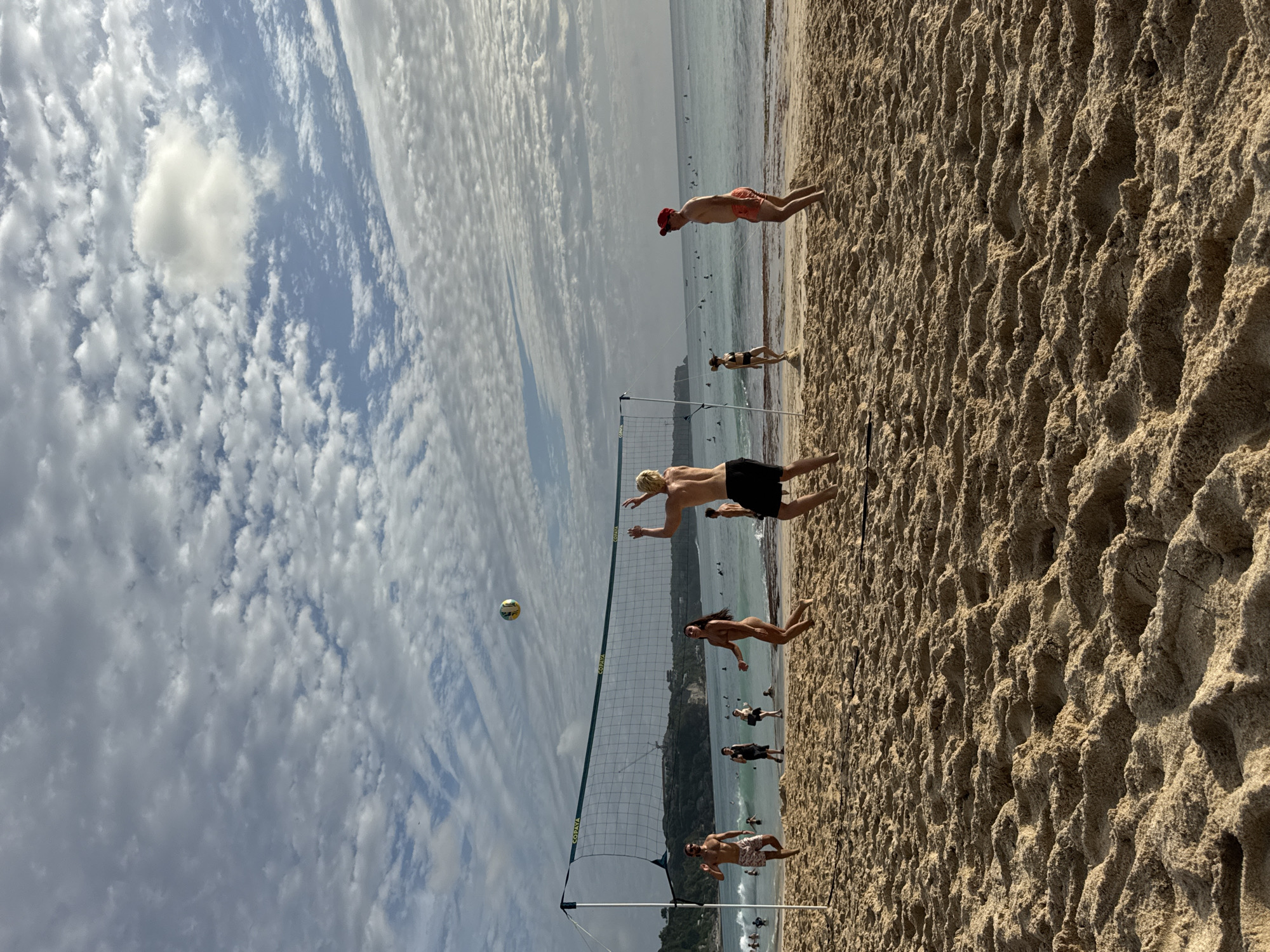The image depicts a beach volleyball game in progress at Kuta Selatan, Indonesia. Several individuals are engaged in the game on a sandy beach, with a volleyball net set up. The ocean is visible in the background, with waves and several people in the water, some appearing to be surfing. The sky is partly cloudy with a mix of white and gray clouds against a blue backdrop. The sand is textured and uneven. The people in the foreground are dressed in swimwear, actively participating in or observing the volleyball match. The overall scene is one of recreation and leisure on a tropical beach.