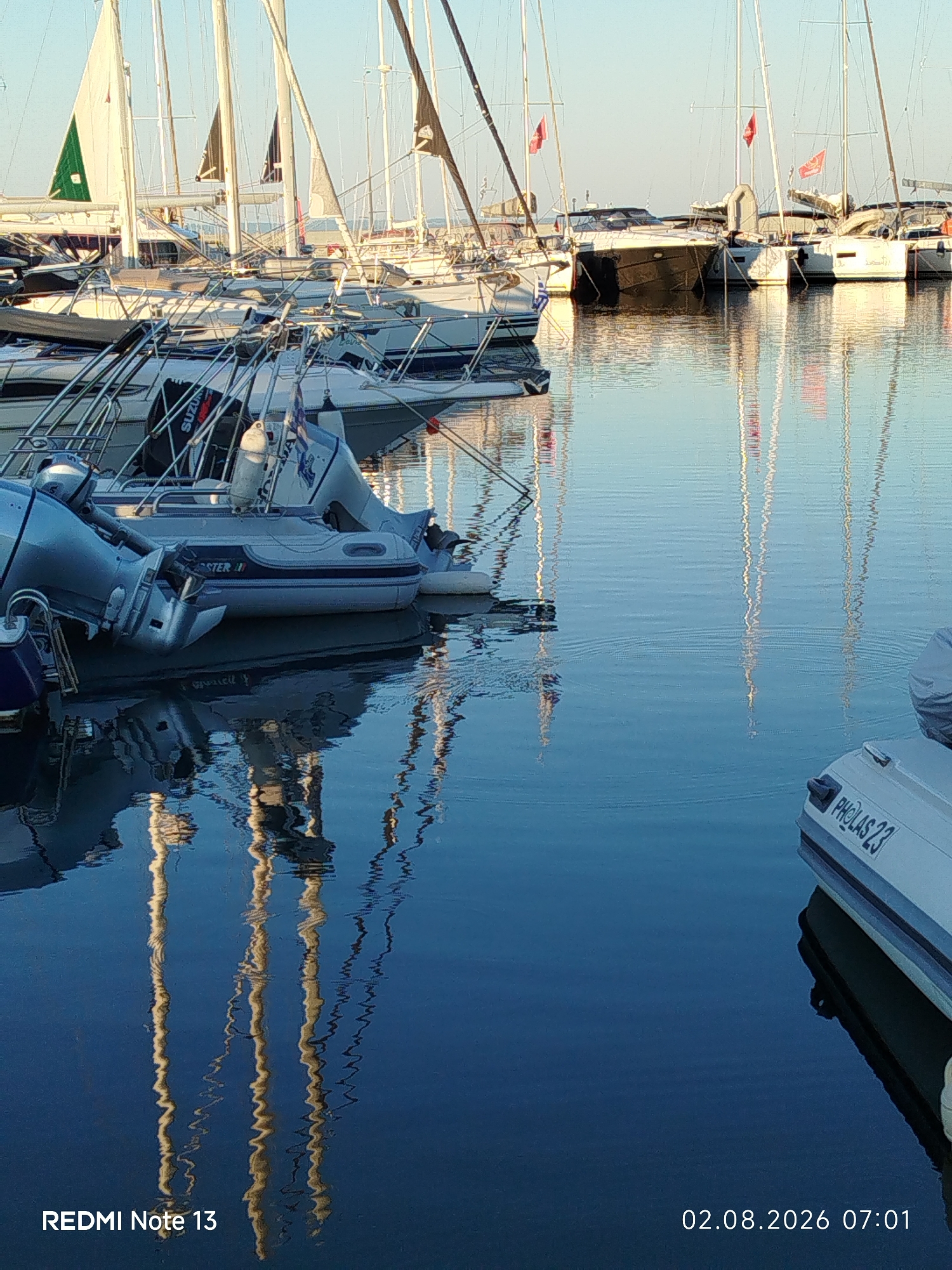 The image depicts a serene marina scene with numerous sailboats and smaller boats docked on calm, reflective water. The boats are white and gray, with masts and rigging clearly visible against a pale blue sky. Some of the boats have red flags. The reflections of the boats and their masts create distorted, wavy patterns on the water's surface. In the foreground, the text "REDMI Note 13" is visible in the bottom left corner, and a date and time stamp "02.08.2026 07:01" is in the bottom right corner. The scene suggests a peaceful morning in a coastal area. This image was captured in the Sithonia Municipal Unit, Greece, a region known for its picturesque coastlines and numerous harbors.