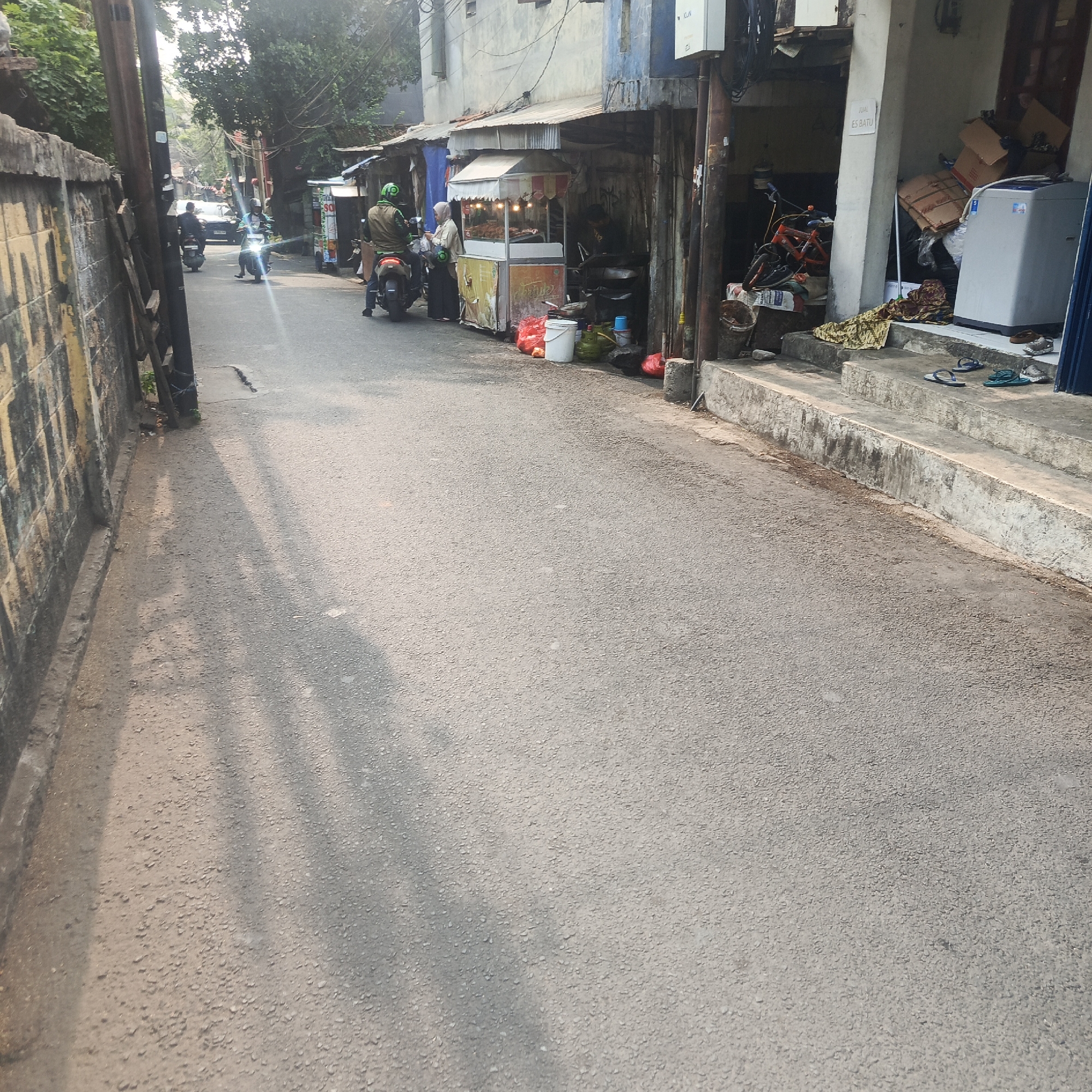 The image depicts a street scene in South Jakarta, Indonesia, characterized by its narrow asphalt road, bordered by buildings and a concrete wall. On the right side of the street, a food stall with a white awning is visible, attended by a person. Several motorcycles are parked along the street, with two riders on motorcycles in motion. One rider, wearing a green helmet, is stopped at the food stall, while another rider in a black helmet and jacket is approaching from the background. The buildings along the street appear to be a mix of residential and commercial establishments, with some displaying signs in Indonesian. Several items are placed on the steps leading up to one of the buildings, including a washing machine and boxes. The overall impression is of a bustling urban environment with everyday activities taking place.